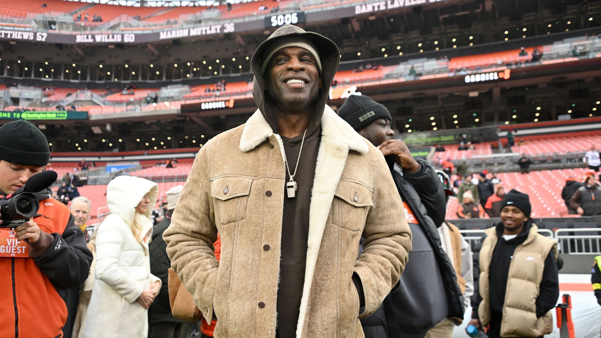 Smiling man wearing brown shearling jacket and hoodie at a sports stadium, with several people in winter clothing and empty red seats in the background.