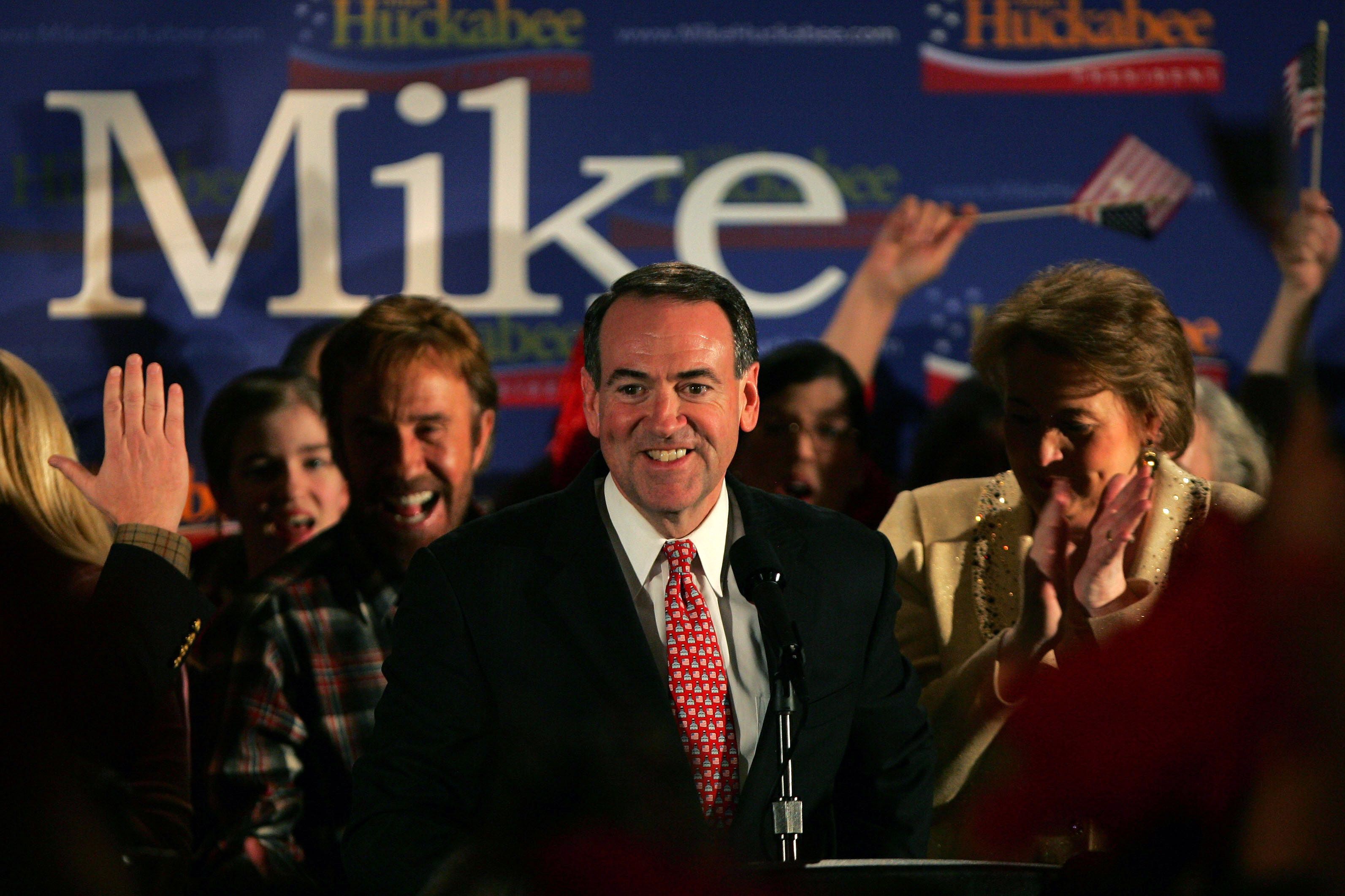 Mike Huckabee smiles with a small crowd arround him. In the background a sign says his first name. 