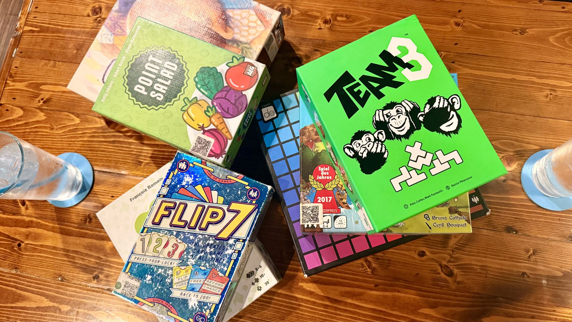 Overhead view of a wooden table covered with colorful board game boxes; a bright green box with bold black lettering and cartoon apes on the right, surrounded by other games with fruits and grid patterns.