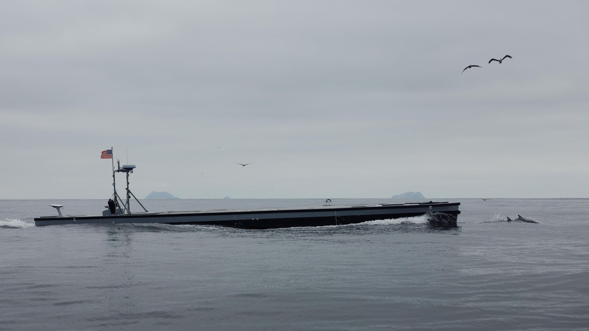 A semisubmersible boat glides through the water. Birds are seen above it. The water and skies are grey.