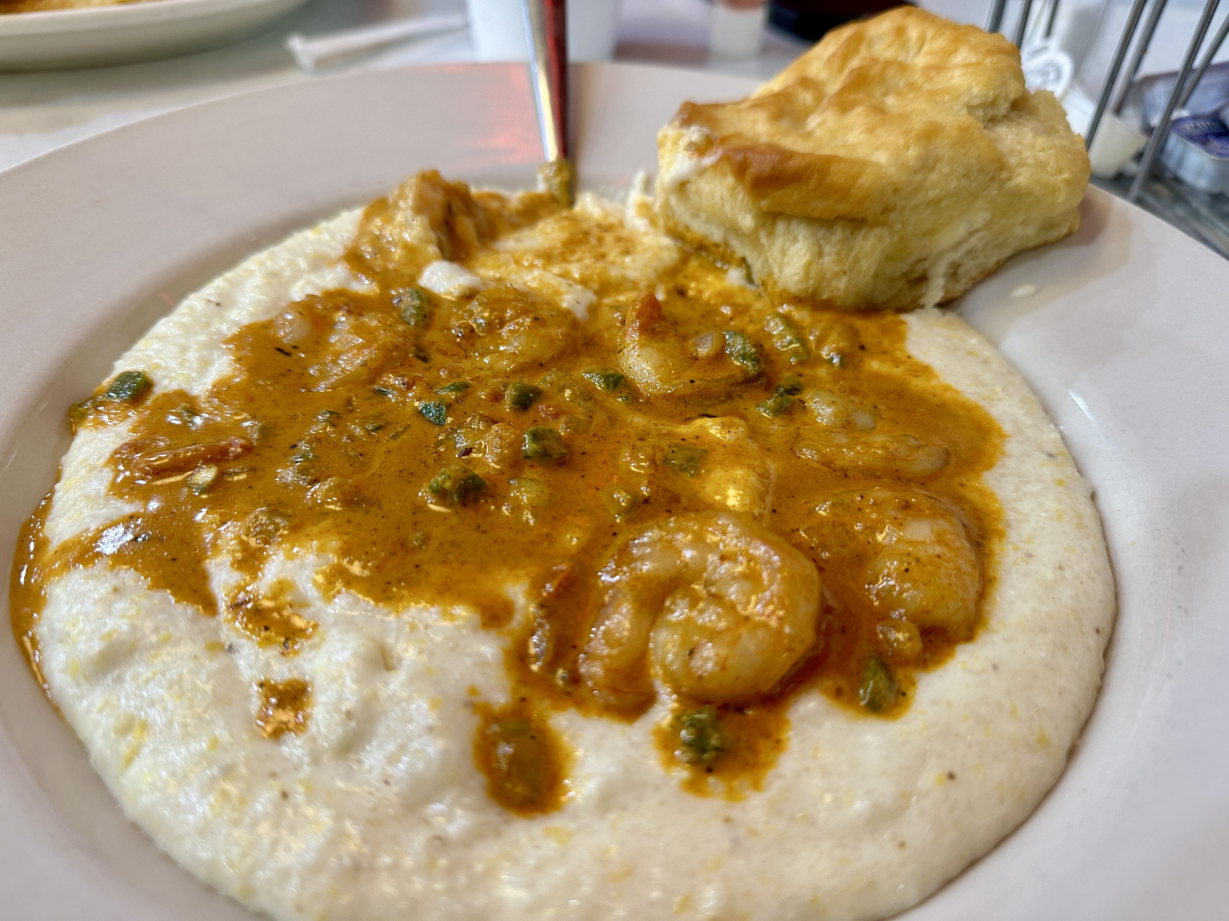Close-up of shrimp and creamy orange sauce served over white grits, accompanied by a golden brown biscuit on a white plate.