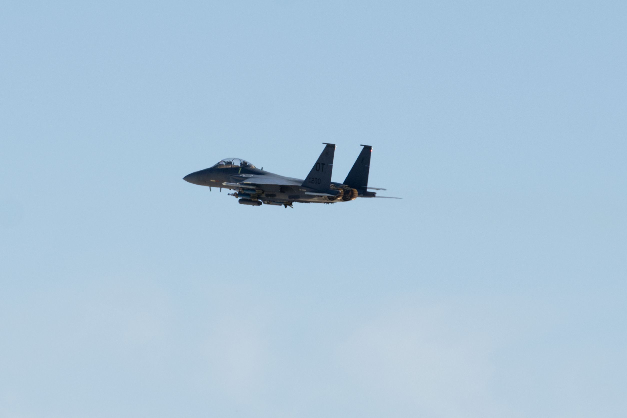 Gray military fighter jet flying in clear blue sky with twin tails and visible armaments beneath wings.