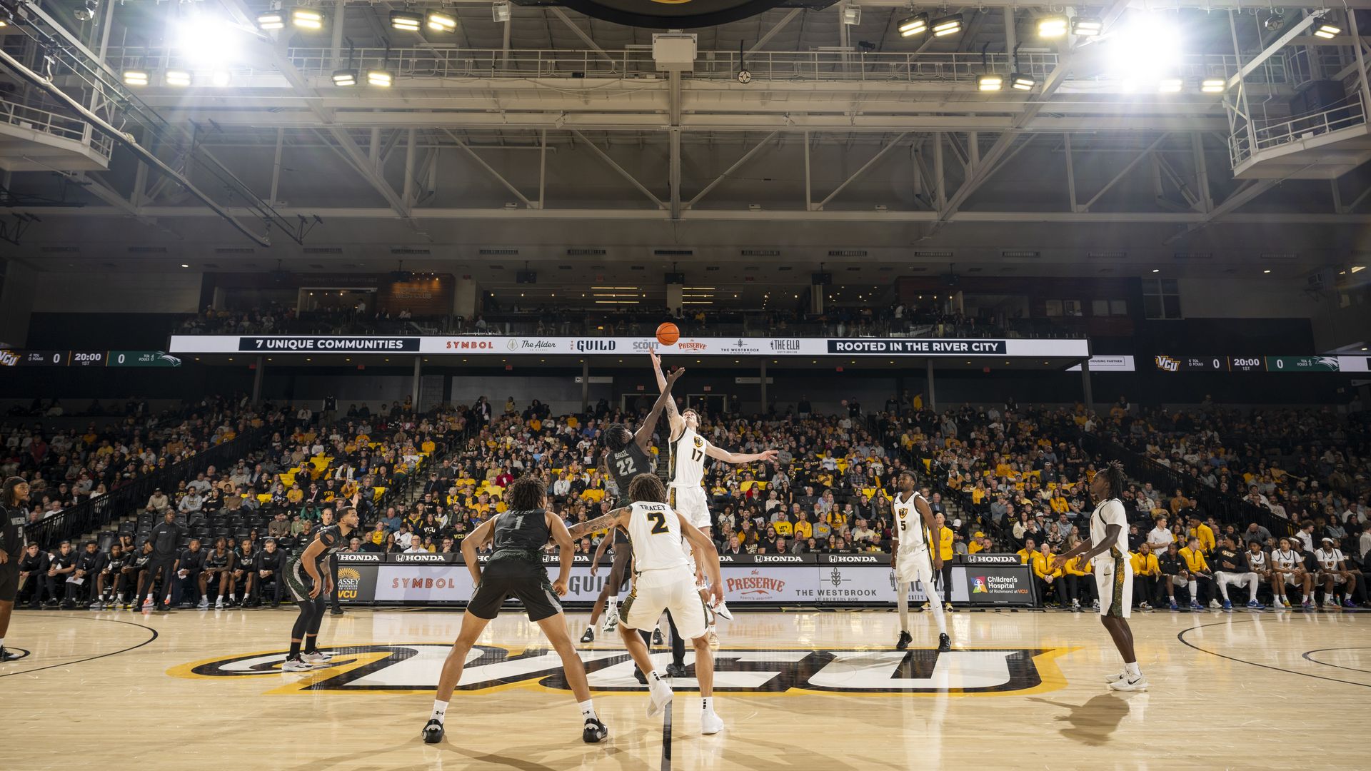 Two basketball players in white and black jerseys jump for the ball during tip-off on a court with large VCU logo, under bright arena lights, crowded stands in background.