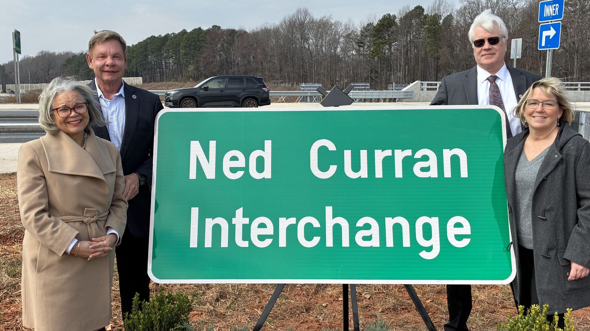 Four people stand outdoors behind a large green road sign reading "Ned Curran Interchange" with plants in front and a road with trees in the background under a gray sky.