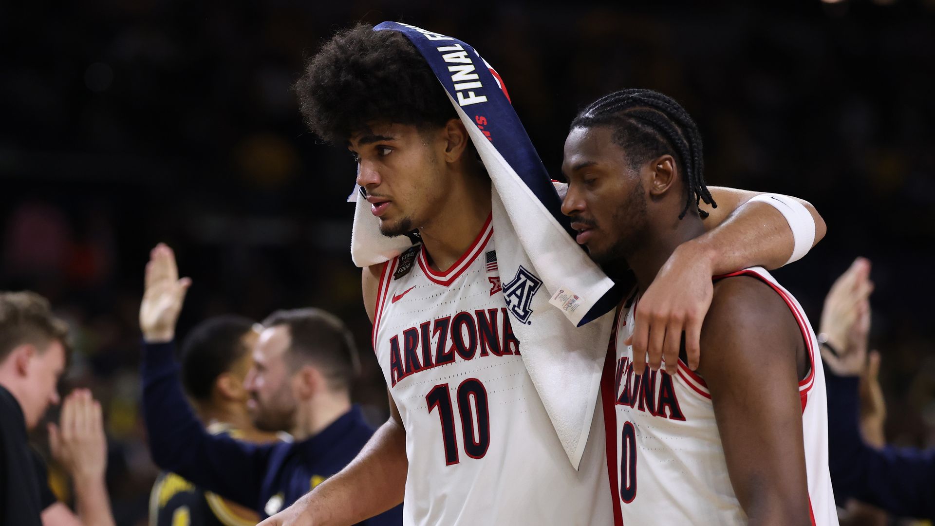 Two basketball players in white University of Arizona jerseys stand on the court. One has a towel over his head and has his arm around his teammate's shoulders. Opposing players celebrate in the background. 