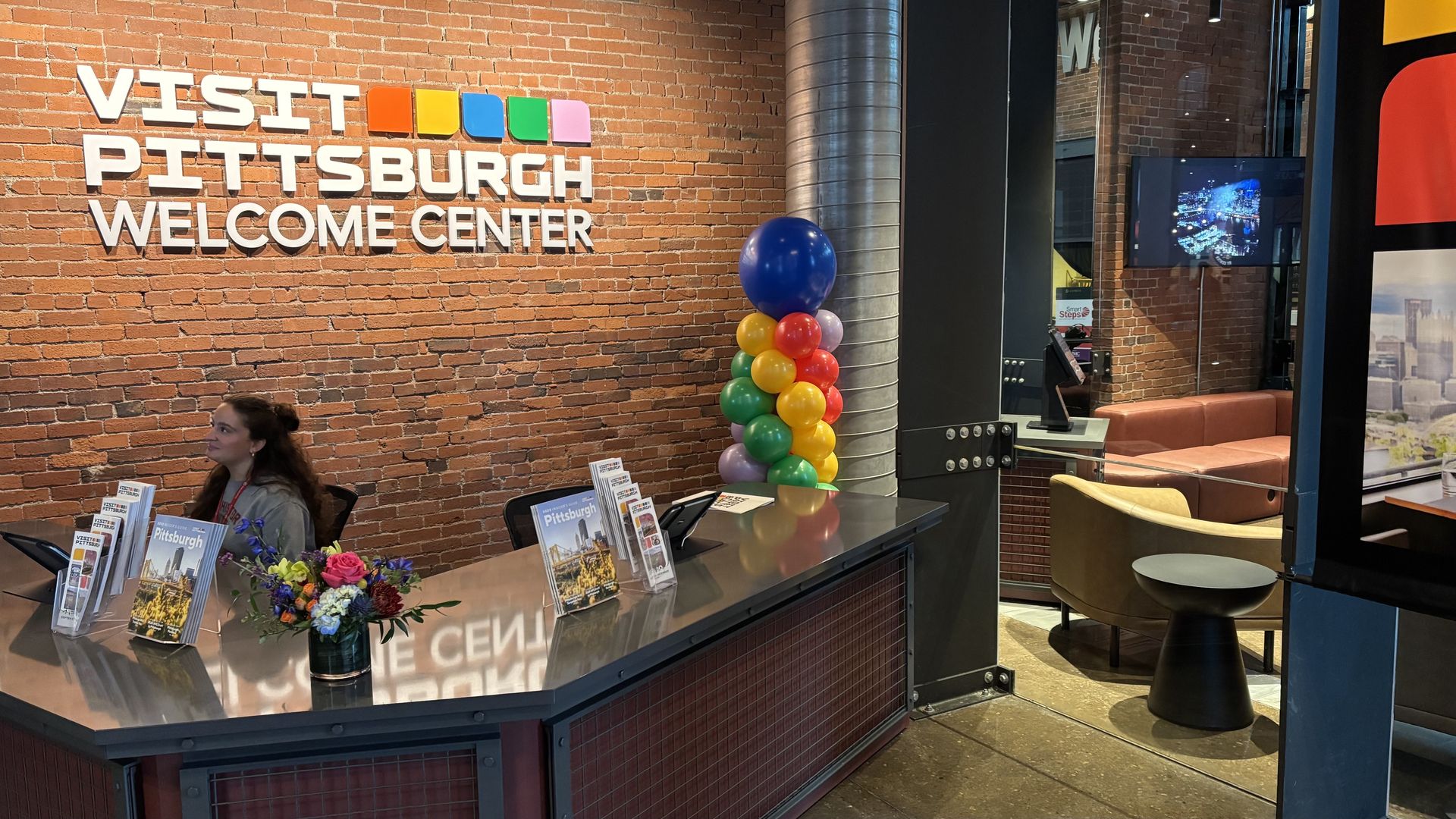 Interior of Visit Pittsburgh Welcome Center with brick wall, colorful logo, balloons, brochures on a steel desk, a woman seated behind it, and a seating area with brown and yellow chairs.