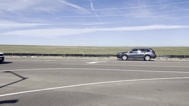 Wide highway scene with a gray SUV in the right lane, a grassy plain and fence in the background, clear blue sky, power lines overhead, white road markings, and another car visible on the left.