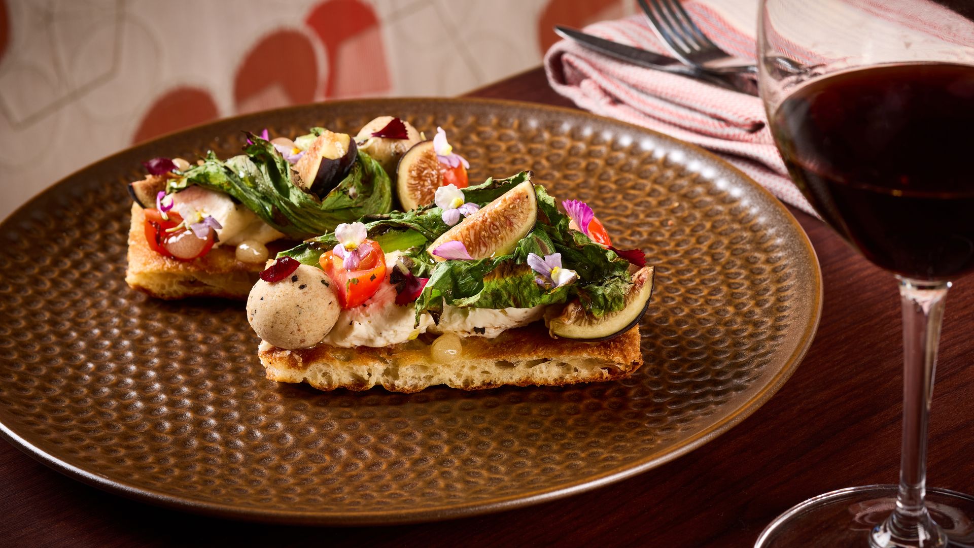 Two crostini topped with greens, figs, cherry tomatoes, edible flowers, and a creamy spread on a textured bronze plate; a glass of red wine and cutlery on a wooden table.