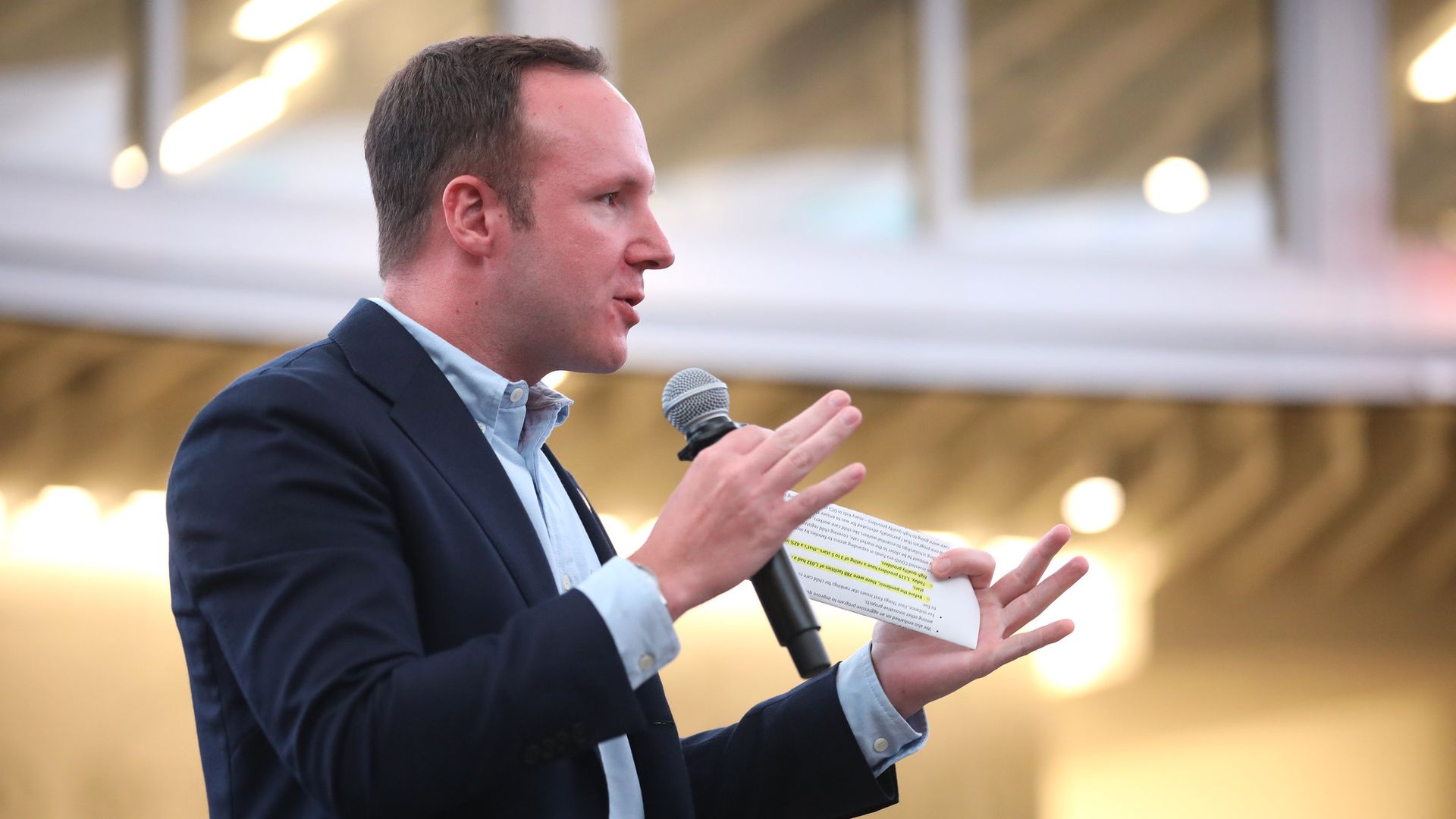 Profile of Matt Gress in a navy suit speaking into a handheld microphone, holding a highlighted sheet of notes, with a blurred, warm-lit indoor background.