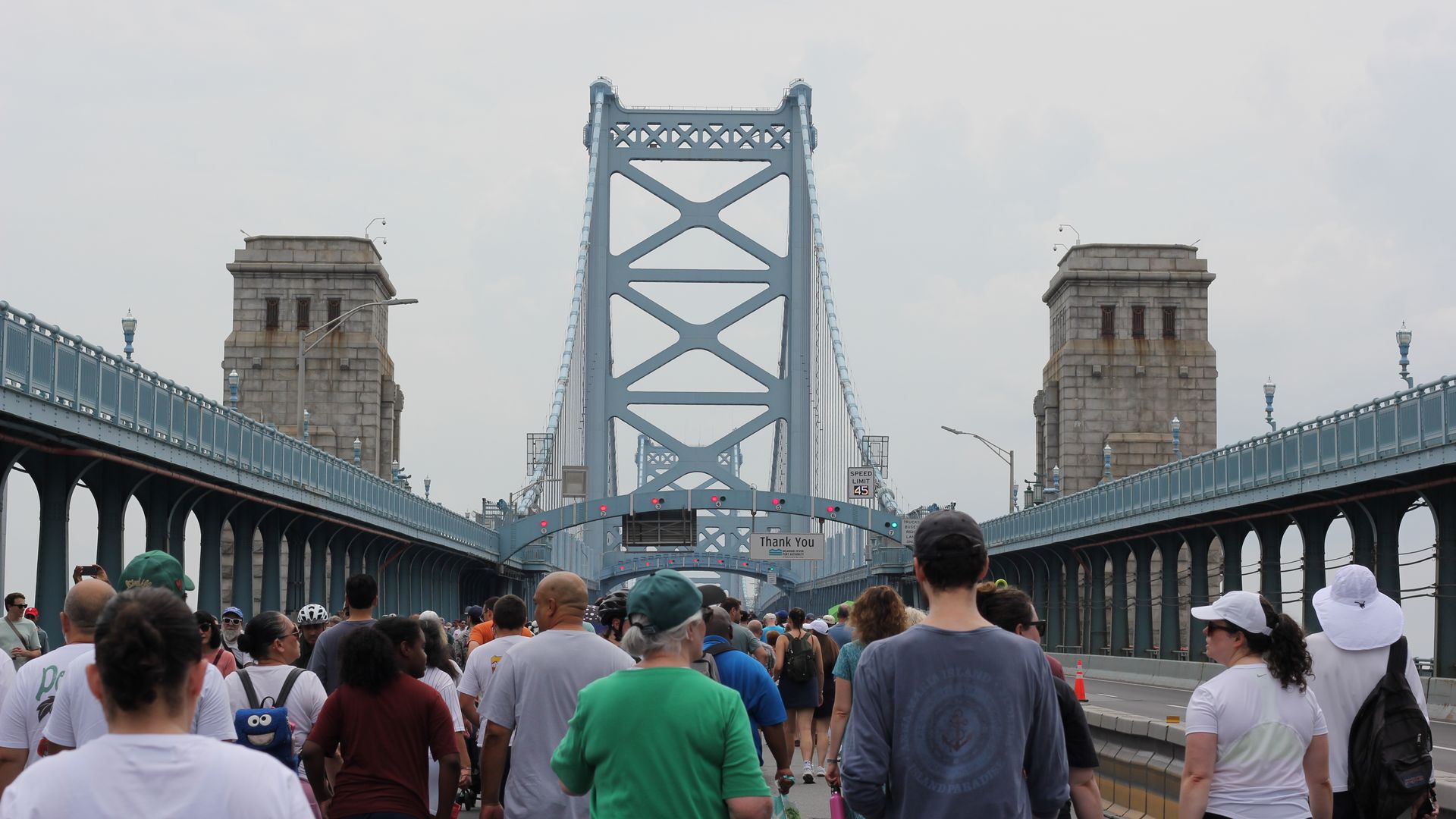 Crowded pedestrian walkway on a pale-blue steel suspension bridge, flanked by stone towers, with many people wearing hats and backpacks under a light sky.