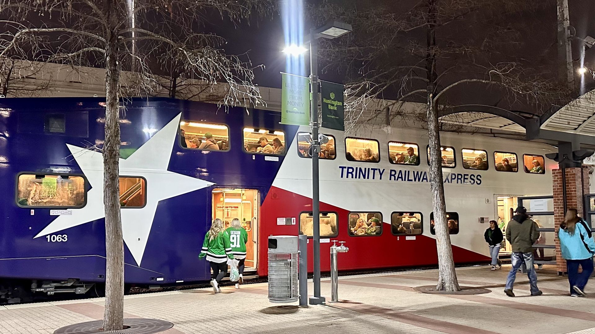 Night scene of a "Trinity Railway Express" double-deck train with a blue, white, and red star livery. Passengers board and sit inside windows; station platform, trees, banners visible.