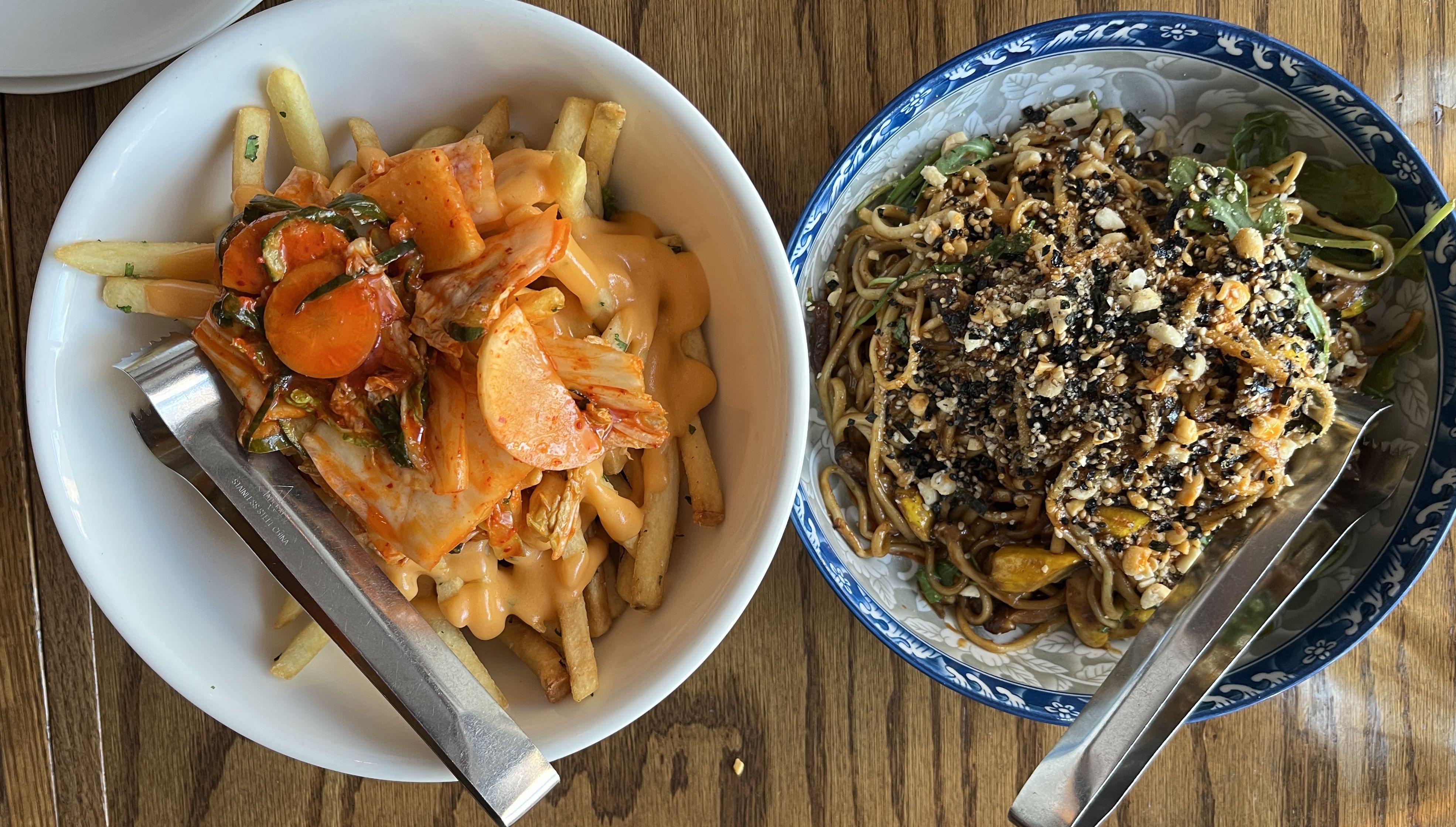 Two bowls on wooden table: left bowl with fries topped with orange sauce and kimchi; right bowl with stir-fried noodles, vegetables, sesame seeds, and crushed nuts in blue patterned bowl.