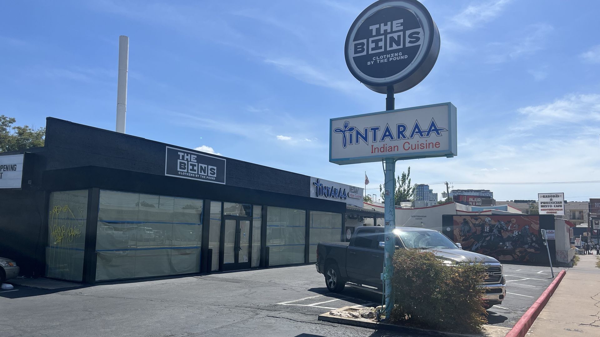 Bright blue sky over a strip mall with a tall circular sign reading "THE BINS" on a pole. A white panel below shows "TINTARAA Indian Cuisine". A dark glass storefront and a gray pickup in the foreground.