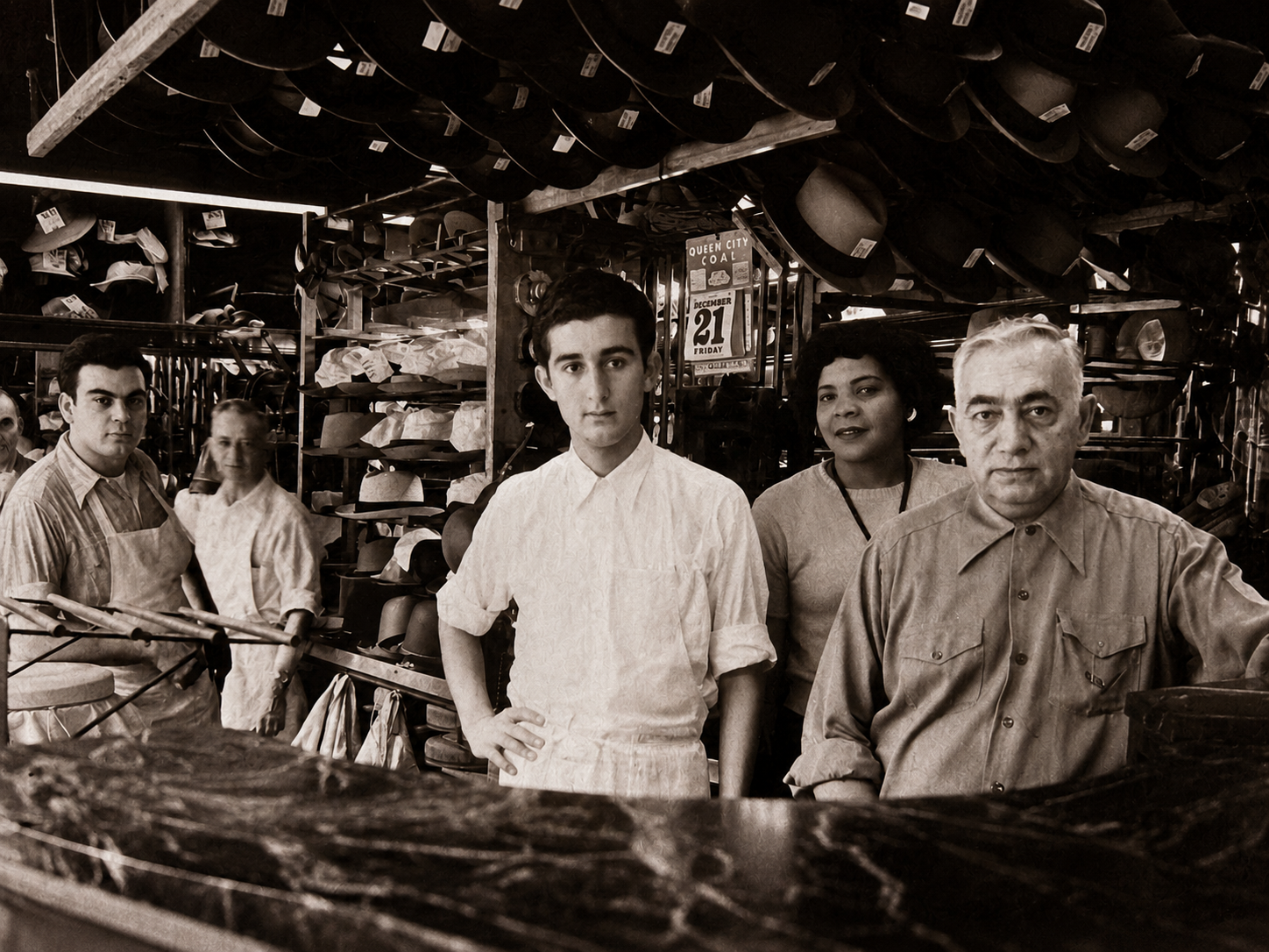 Sepia-toned vintage hat shop with five workers behind a marble counter. Hats line shelves and ceiling racks. Center: a young man in a white shirt, flanked by a woman and an older man.
