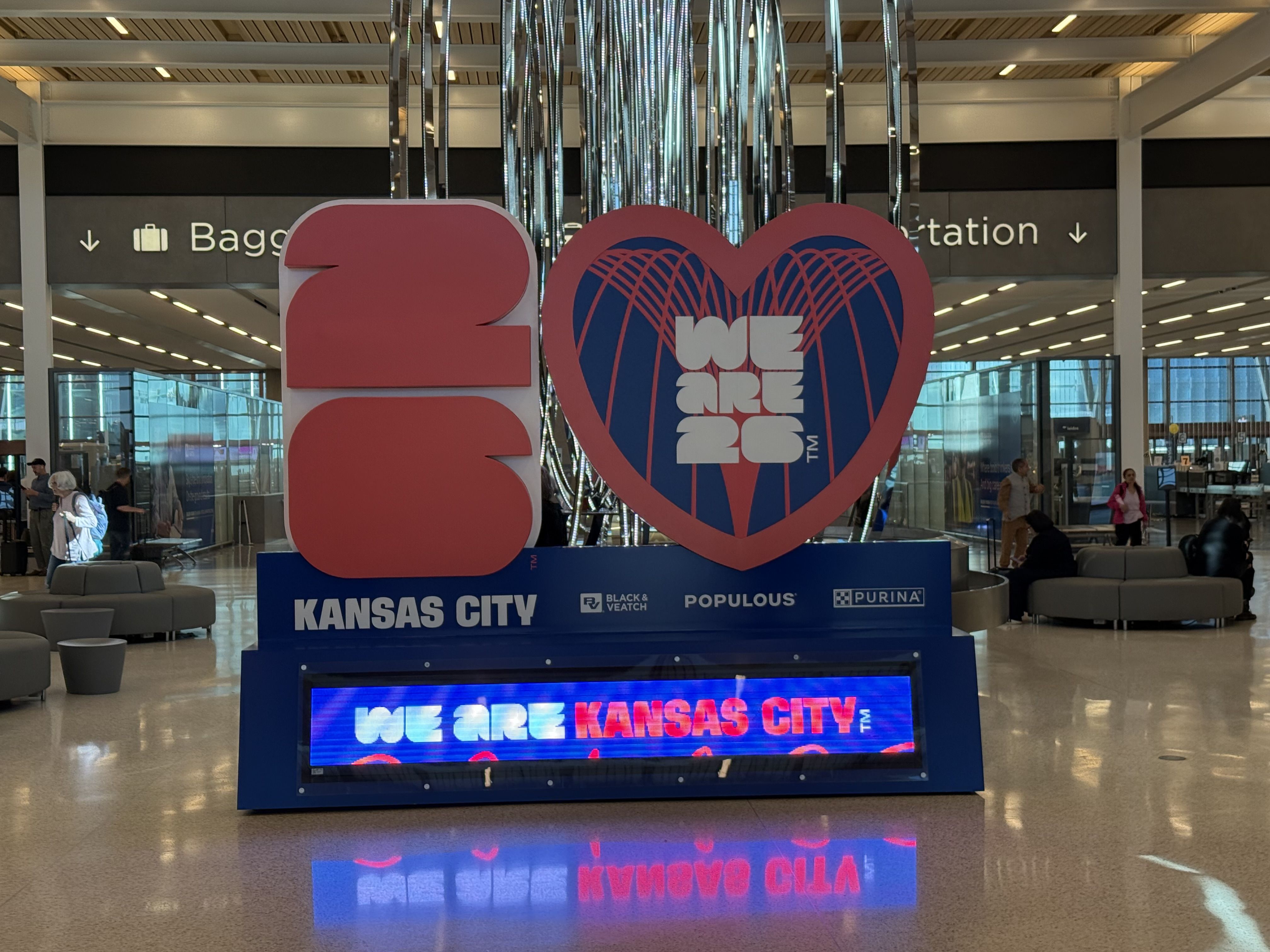 Red and blue 2026 Kansas City display with heart shape in airport terminal showing "We are 26" inside heart, signage above for baggage claim, people sitting and walking nearby.