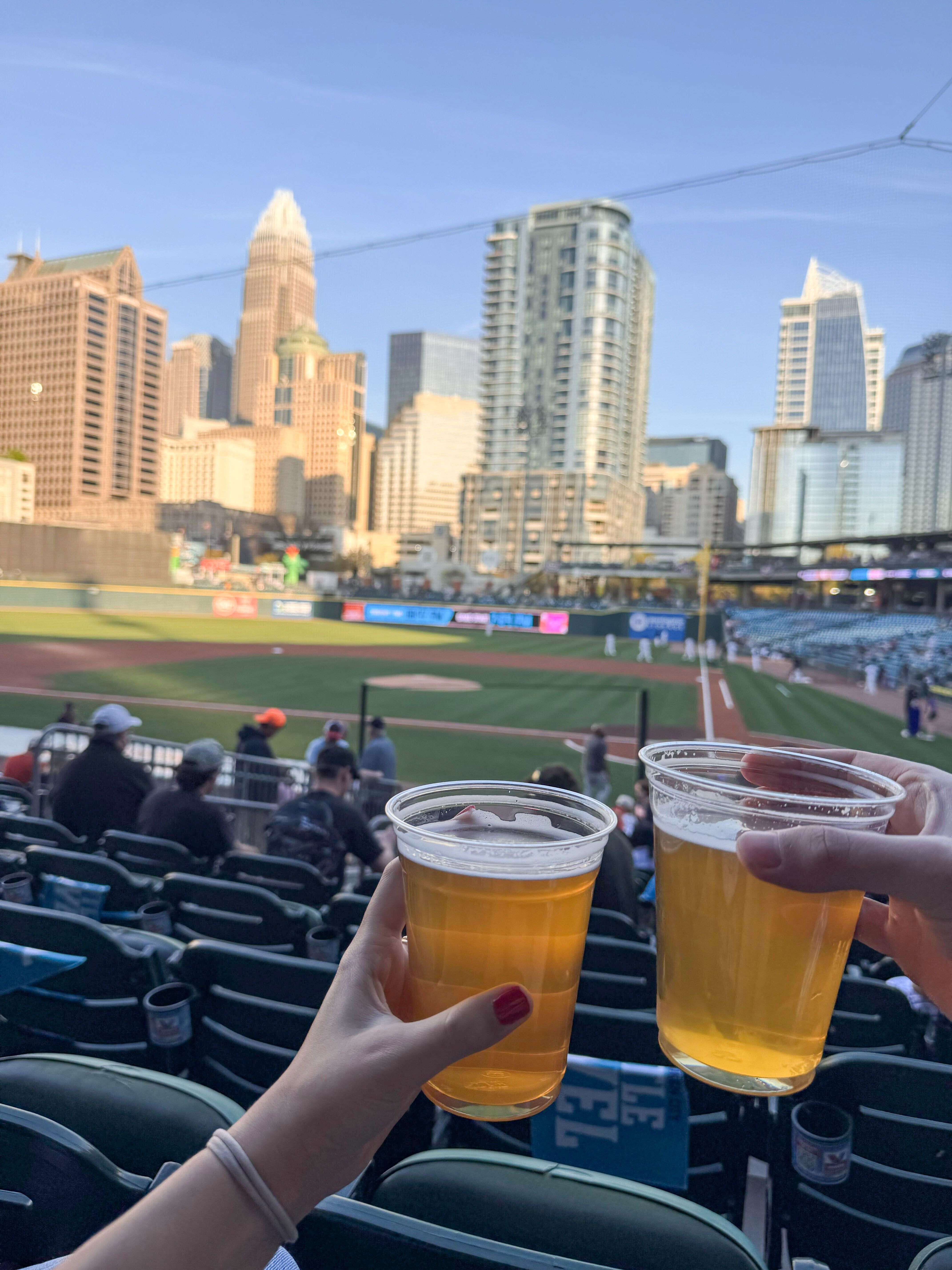 Two people toast with plastic cups of beer against a bright, clear view of the baseball field and city skyline.