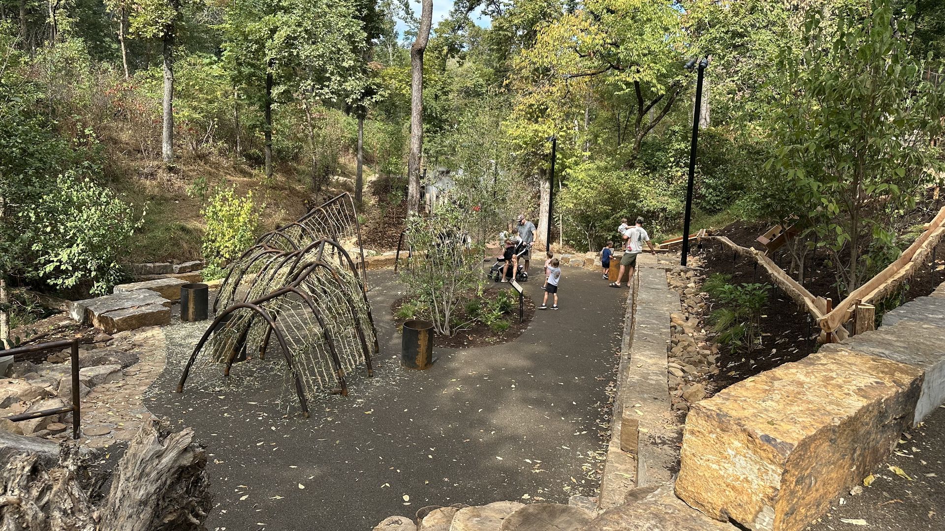 Panoramic view of a wooded park path with stone walls and curved metal railings. A group of people and children stand near the center, surrounded by tall trees and green foliage.