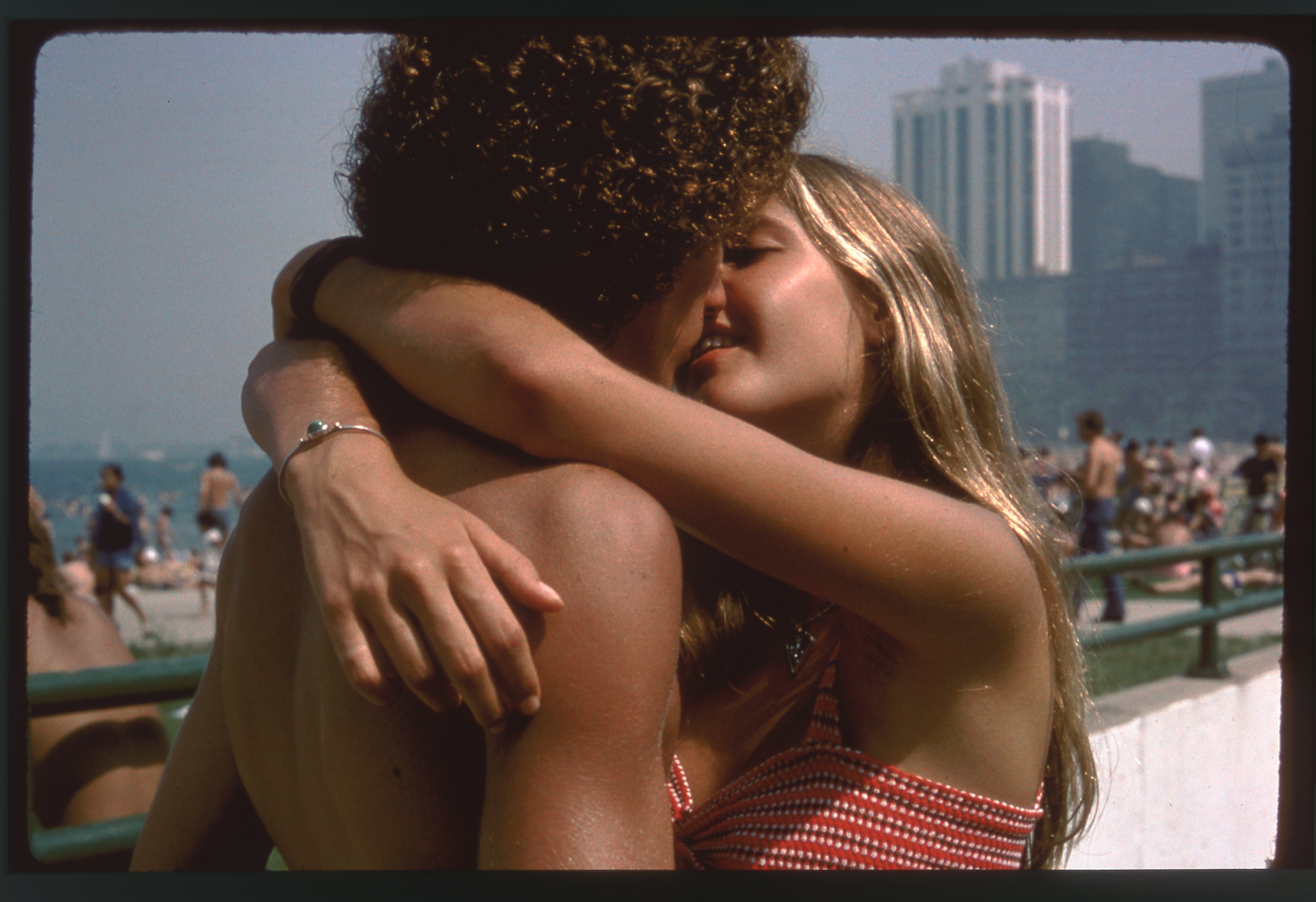 A couple hugs at the beach: a man with curly dark hair and a woman in a red striped bikini, smiling as they embrace; background shows a boardwalk and tall city buildings.