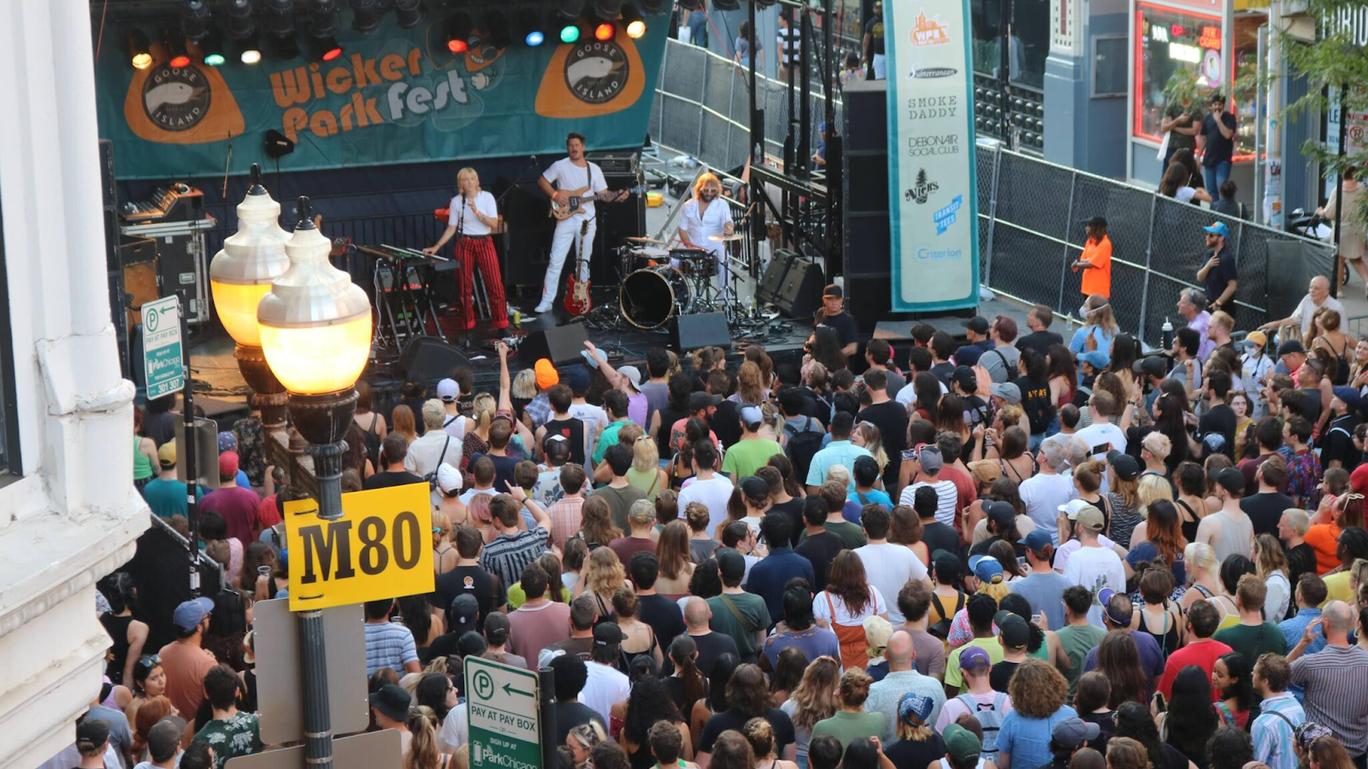 Crowd watching a live band perform on an outdoor stage at Wicker Park Fest with colorful lights and street signs labeled M80 and ParkChicago visible.