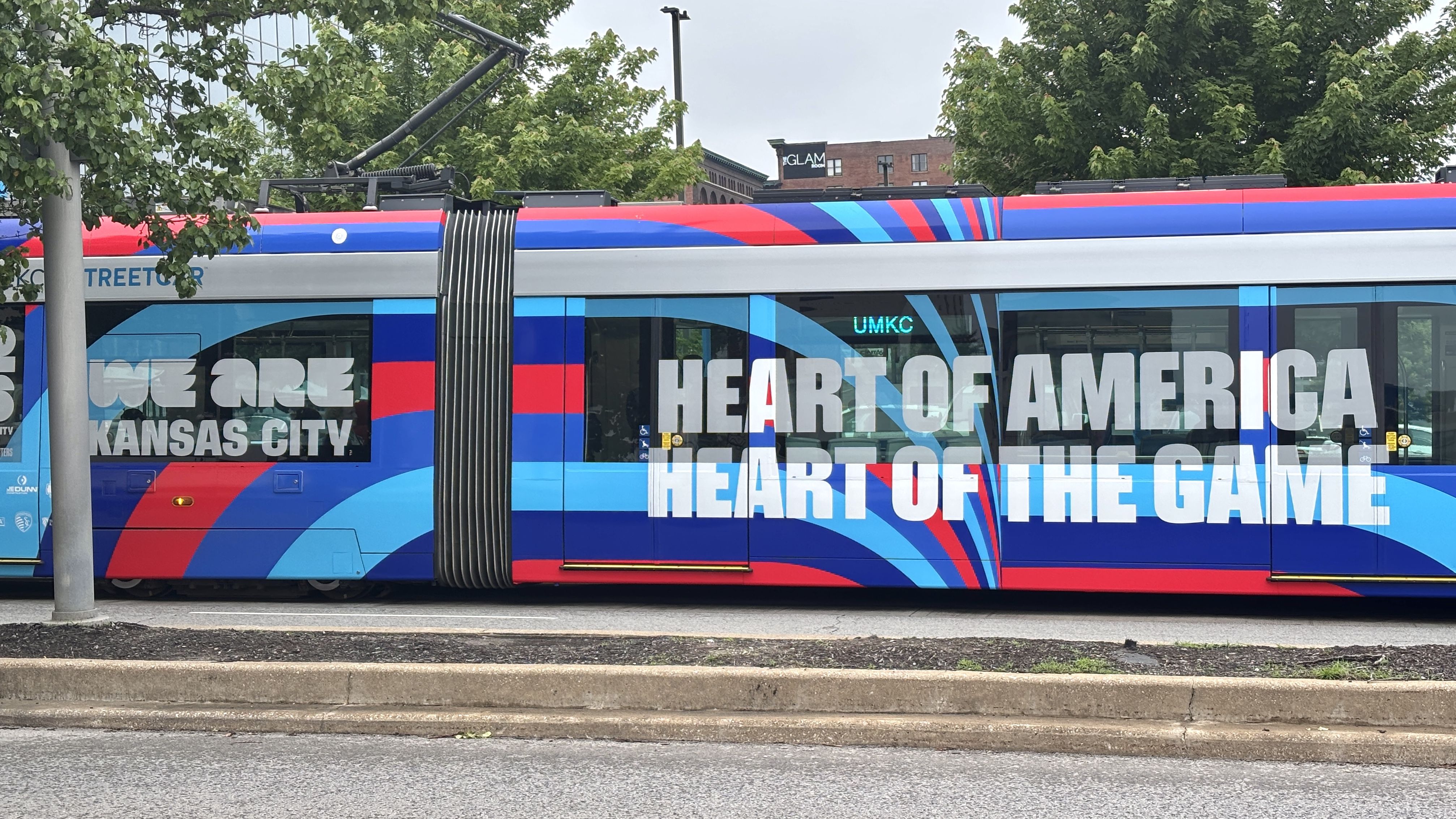 Colorful tram in red, blue, and cyan with large white text reading "WE ARE KANSAS CITY" and "HEART OF AMERICA"; green trees line the street and buildings visible in the background.
