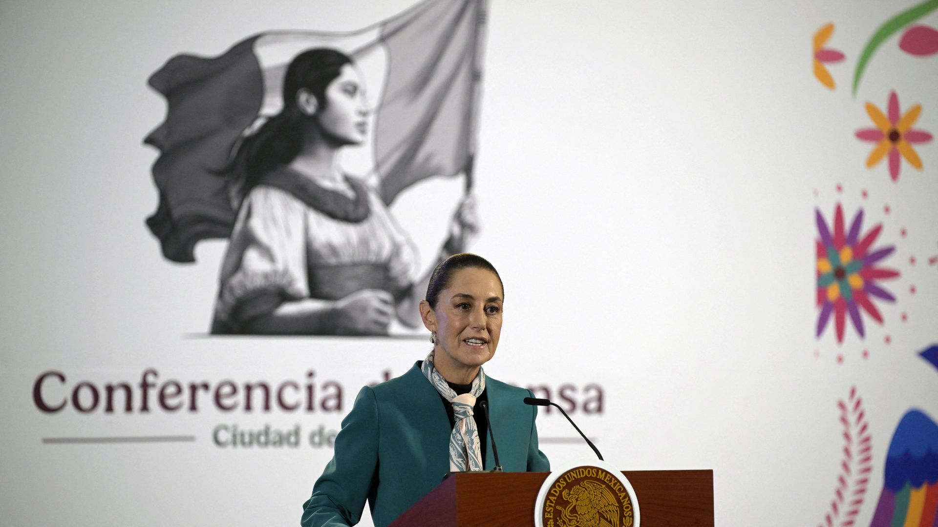 Mexican President Claudia Sheinbaum stands in front of a brown podium, behind her is an illustration of a woman holding the Mexican flag