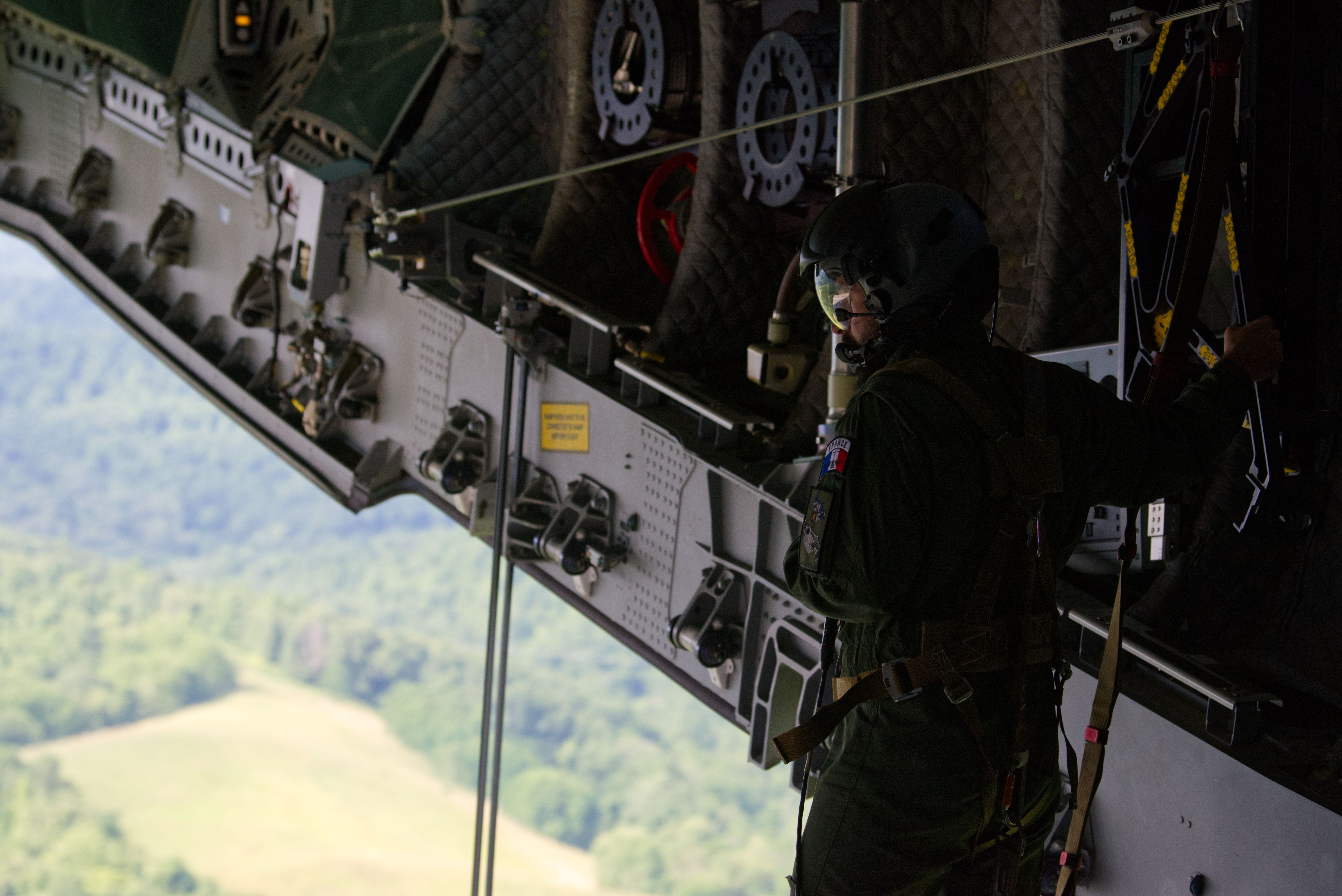 A man in a dark flight suit and helmet stands near the rear ramp of an aircraft as it flys over fields. He's surrounded by gears and pulleys and wires.