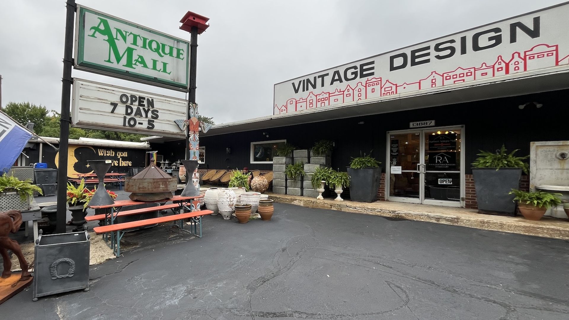 Outdoor view of Antique Mall and Vintage Design store with black facade, potted plants, a red bench, clay pots, an open flag, and a sign showing hours as 10-5, 7 days a week.