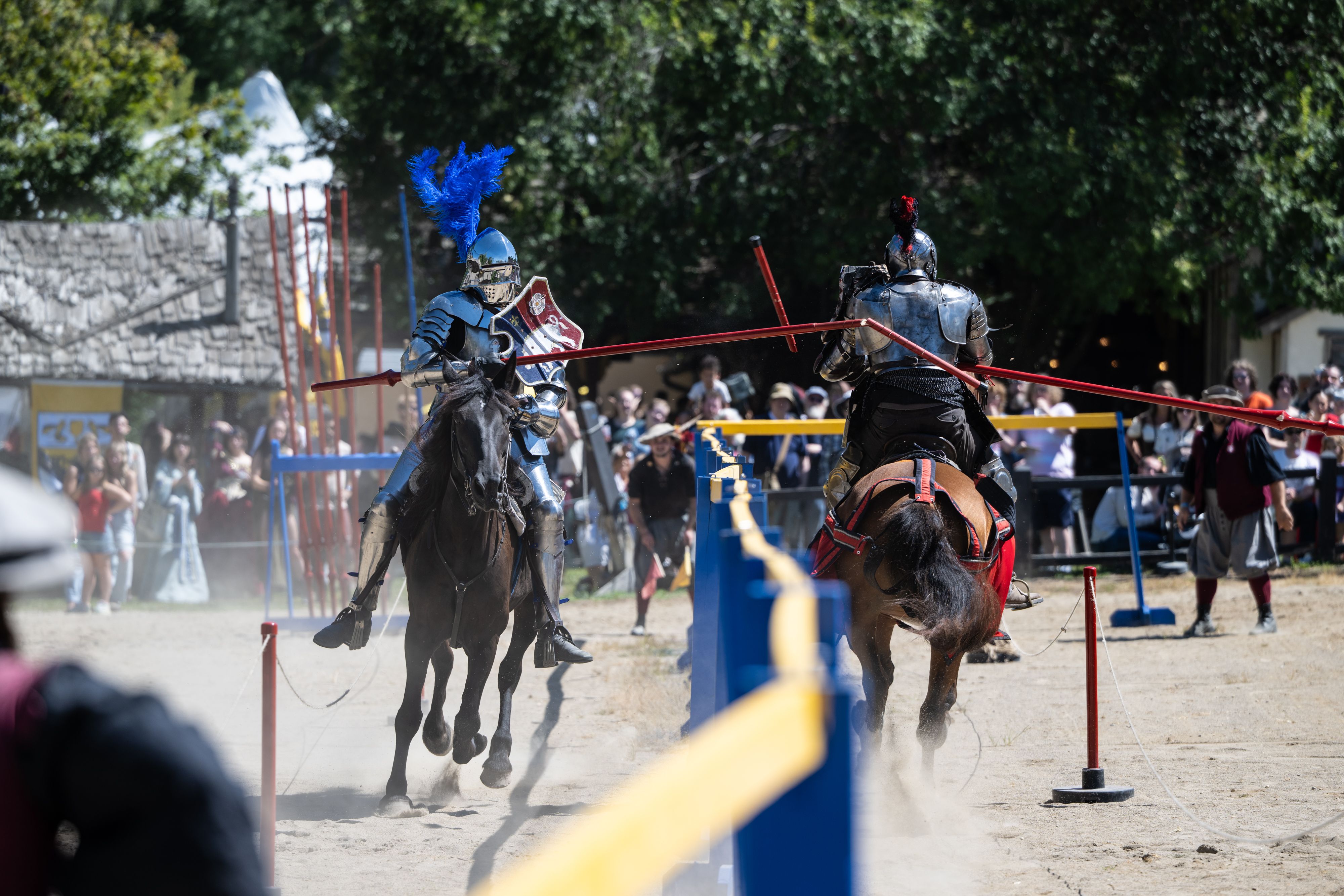 Two armored knights on horseback clash in a jousting ring, one with a blue plume and shield and the other with a red plume. Spectators stand behind yellow and blue barriers.