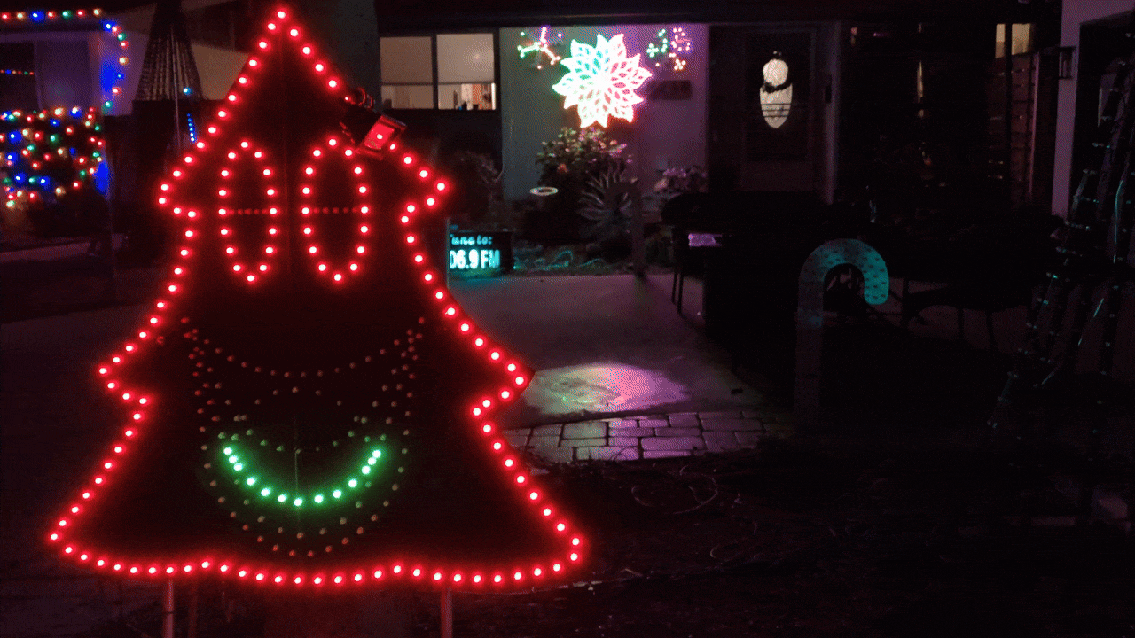 Outdoor Christmas lights display at night featuring a red tree-shaped figure with eyes and a green smiling mouth, a glowing white snowflake on a house, and a lighted candy cane.