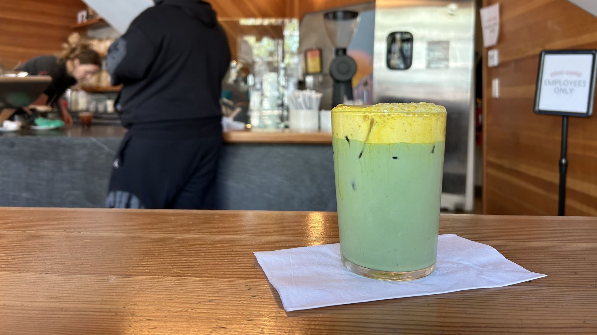 Green layered iced drink with a yellow foamy top on a wooden table with a napkin. Blurred background shows a bar, a worker in black, stainless equipment, and an "Employees Only" sign.
