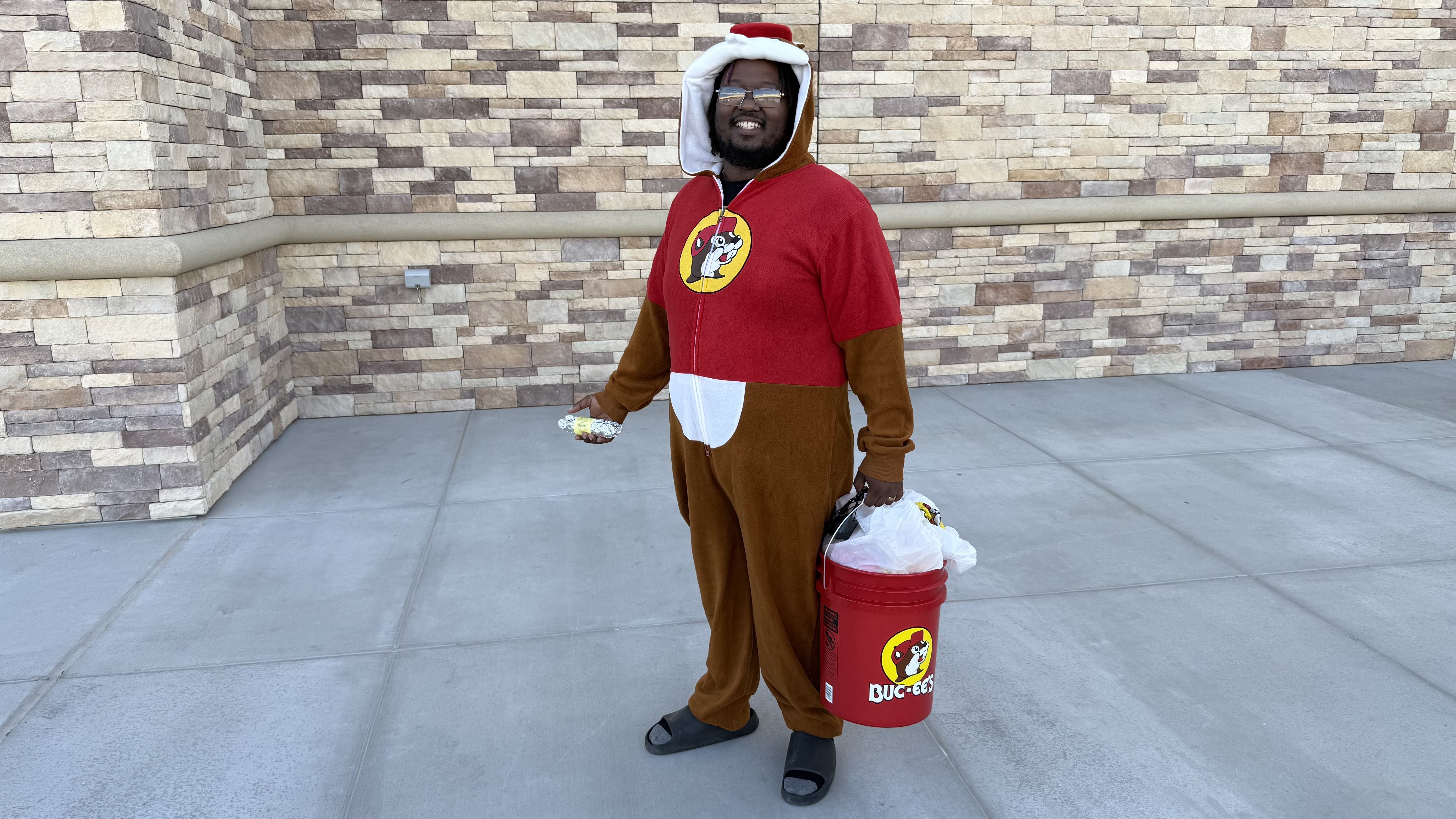A person in a red and brown beaver onesie with a white hood stands on a concrete area in front of a light brown stone wall, smiling, holding foil-wrapped taco and a red Buc-ee's bucket.