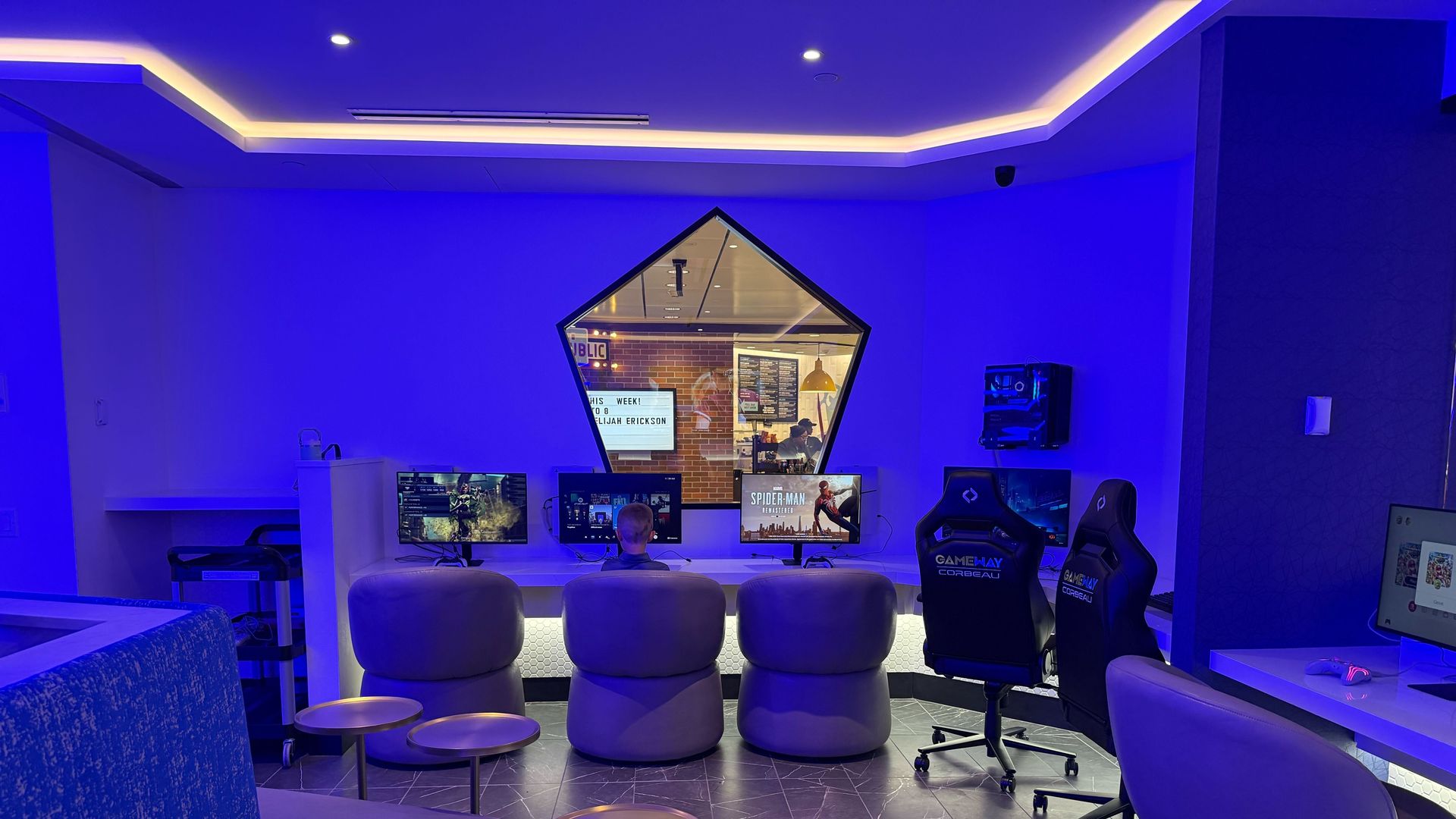 Blue-lit esports lounge with a curved sofa, row of desks and monitors, gaming chairs, and small round stools. A hexagonal window shows a brick-walled room with people at computers.