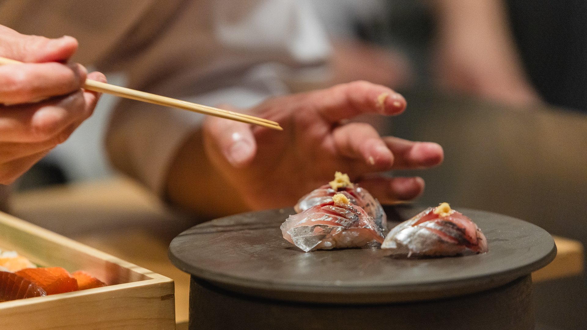 Close-up of hands using chopsticks to pick up nigiri sushi from a dark plate; several fish-topped rice pieces with a wooden sashimi box visible in the foreground.