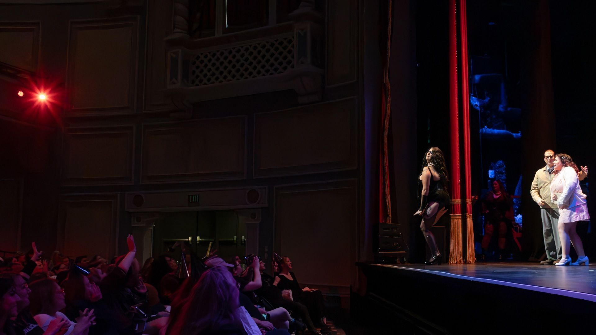 Dark theater stage with a red spotlight on the left and blue lighting on the right. On stage, a few performers stand near a red curtain; audience fills the foreground, many hands raised.
