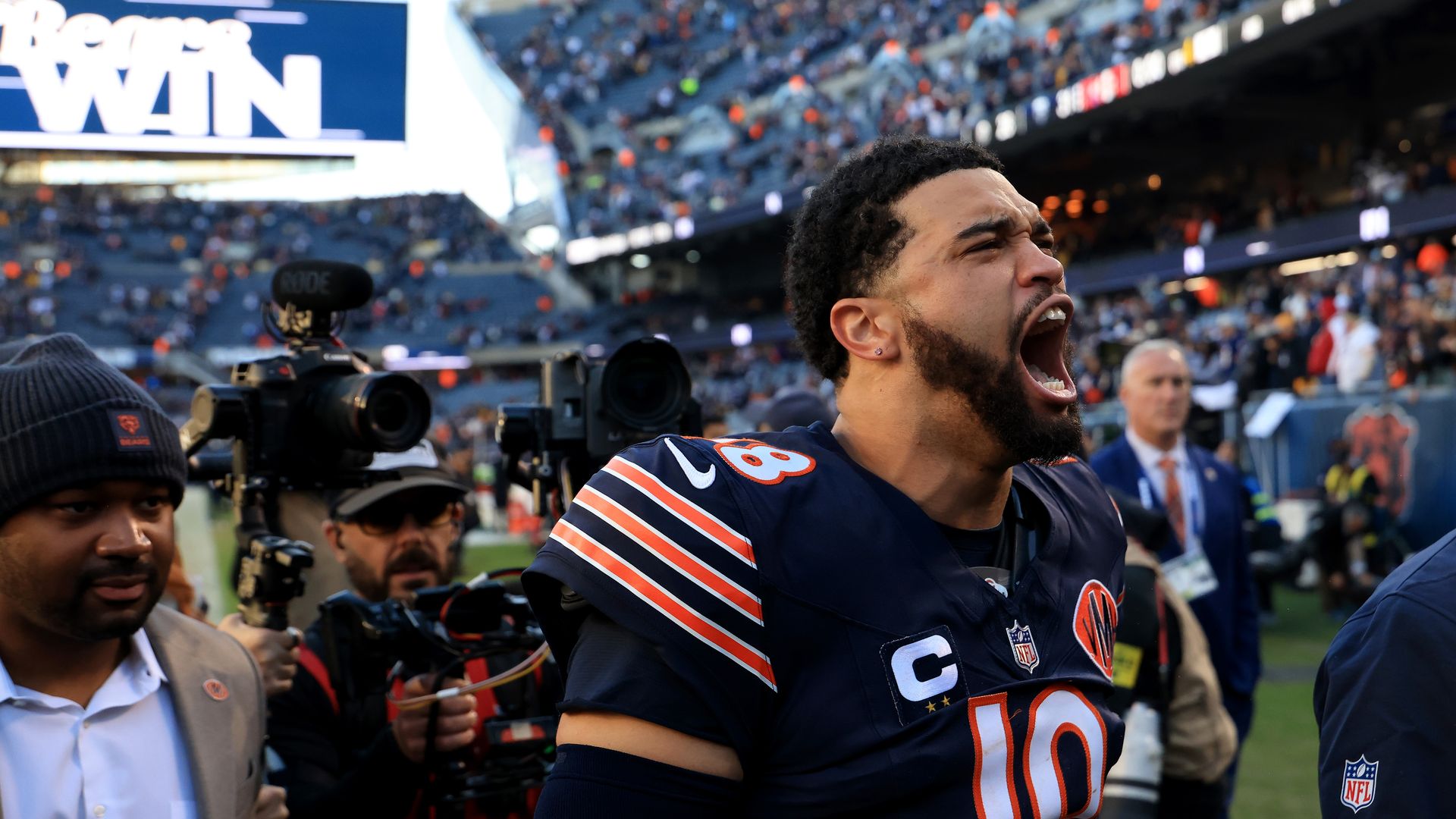 Chicago Bears player #18 in a navy uniform shouting on the field with stadium crowd and cameras behind him. Sign displays a partial message ending with WIN.