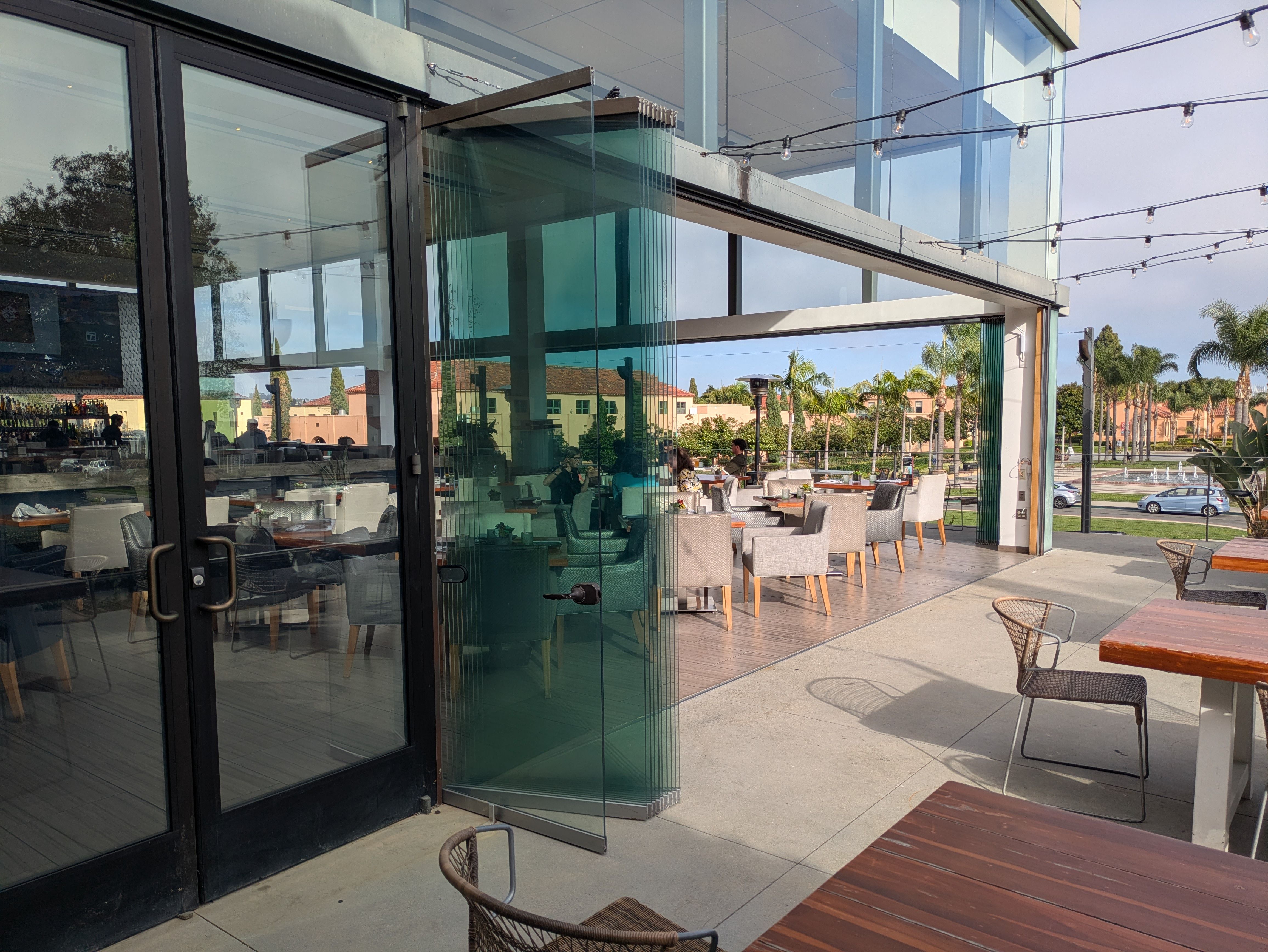 Outdoor cafe with glass doors and walls, string lights, and a covered patio. Wooden tables and grey wicker chairs; patrons dine inside and outside as palm trees line a sunny street.