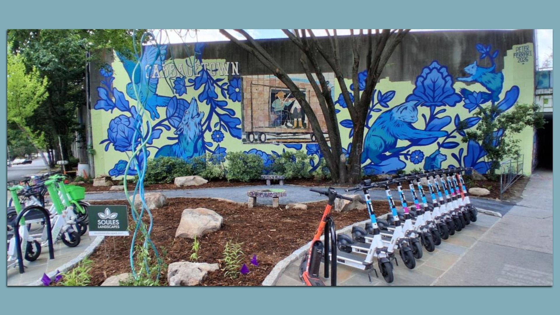 Vibrant mural on a lime wall with large blue flowers and animals, a central inset scene, and foreground trees, rocks, and a line of parked white-and-blue electric scooters in an urban setting.