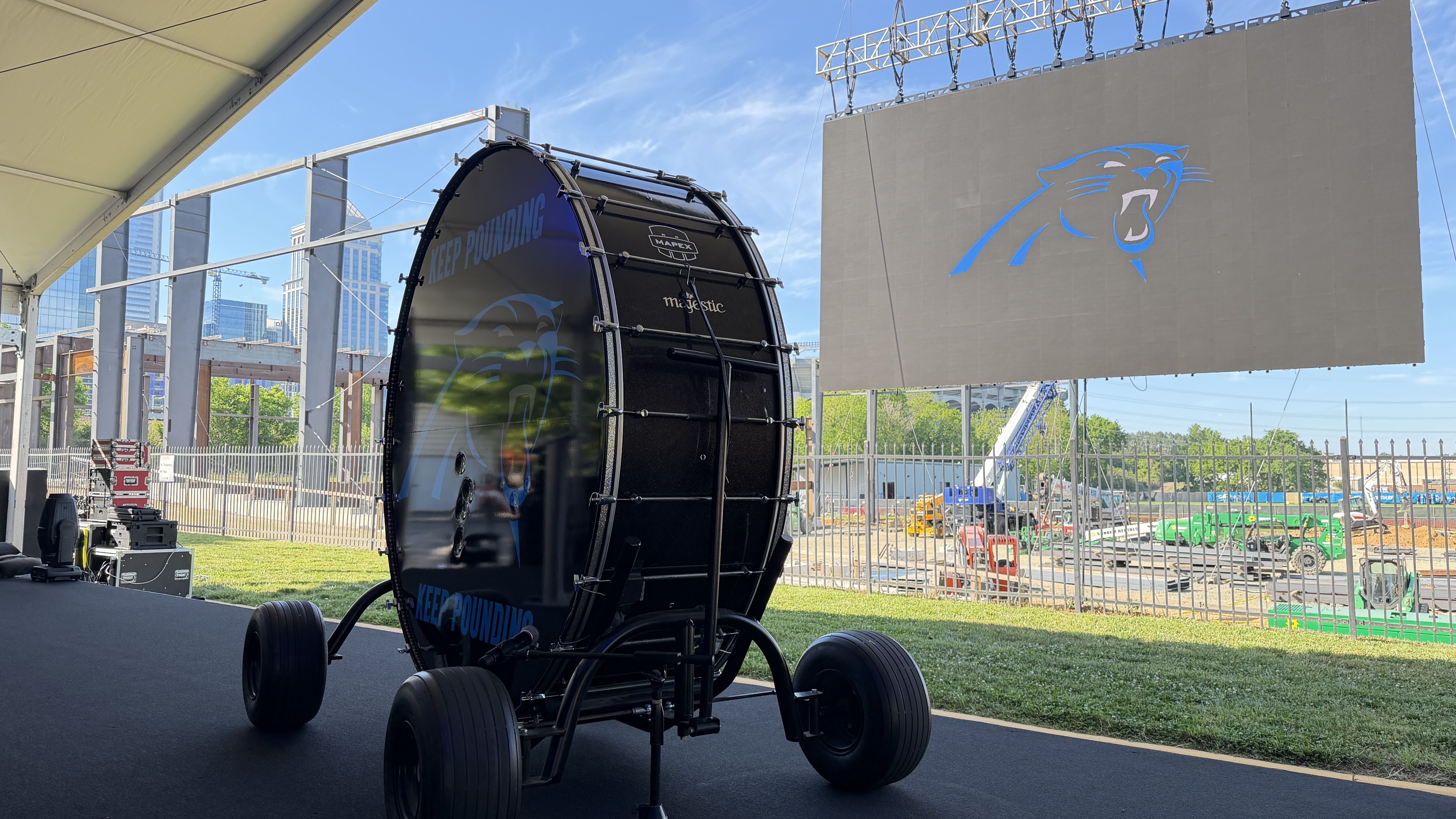 A large black bass drum on a wheeled stand under a tent, with 'KEEP POUNDING' on its side, beside a big screen displaying a blue panther logo; a construction site and city skyline visible in the background.