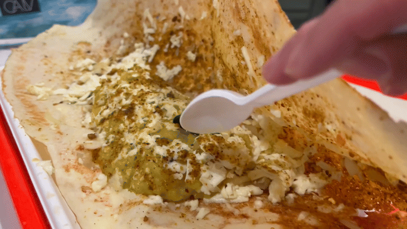 A close-up of a hand using a white plastic spoon to spread green and white chutney or sauce with spices over a large brown dosa on a red tray.