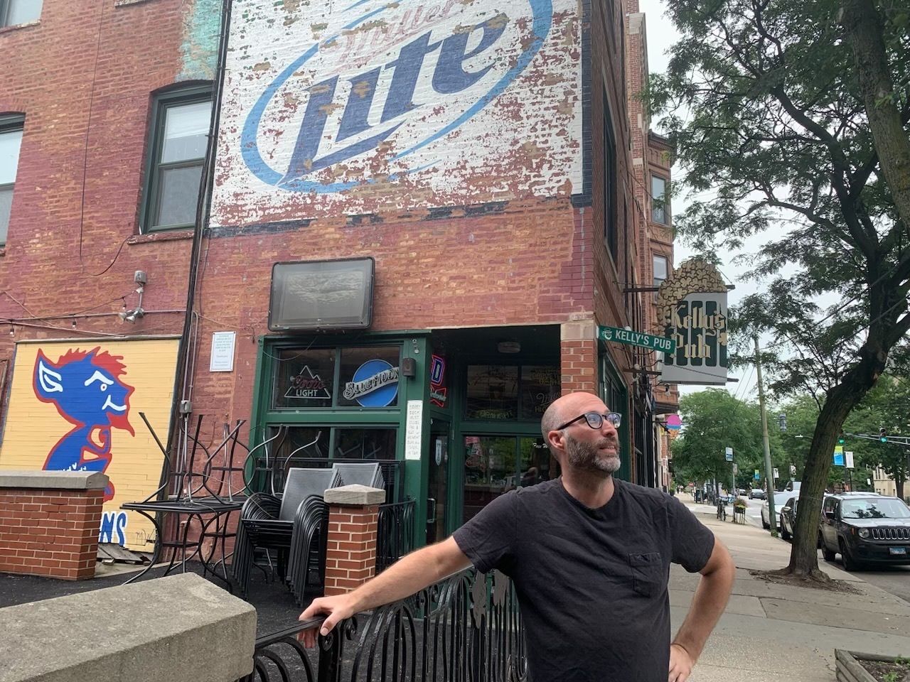 Man in black shirt standing outside a red brick building that has a faded Miller Lite sign.