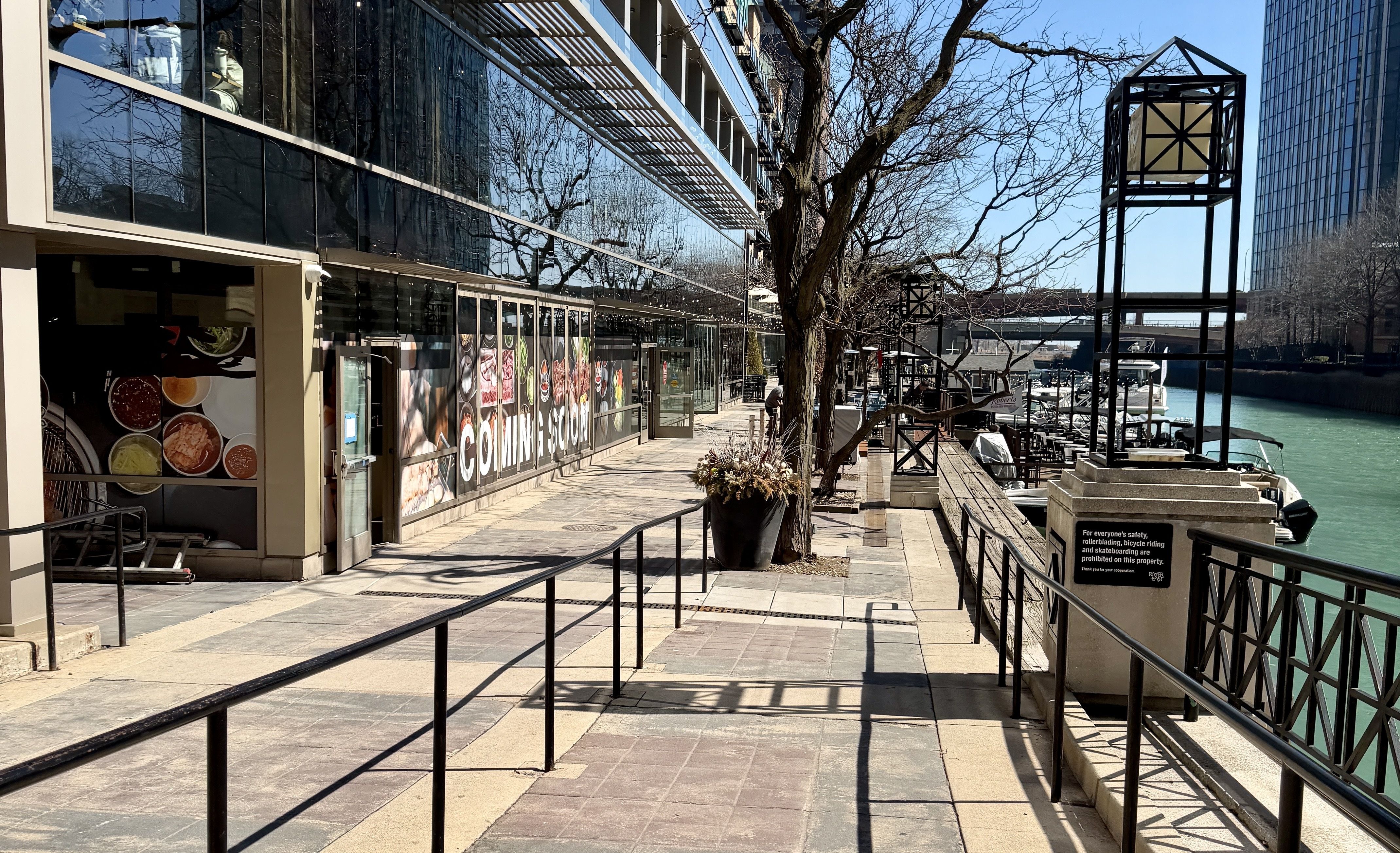 Sunny riverside promenade beside a glass building with a large "COMING SOON" sign on the windows. Bare trees line the walkway; turquoise water with docked boats to the right.
