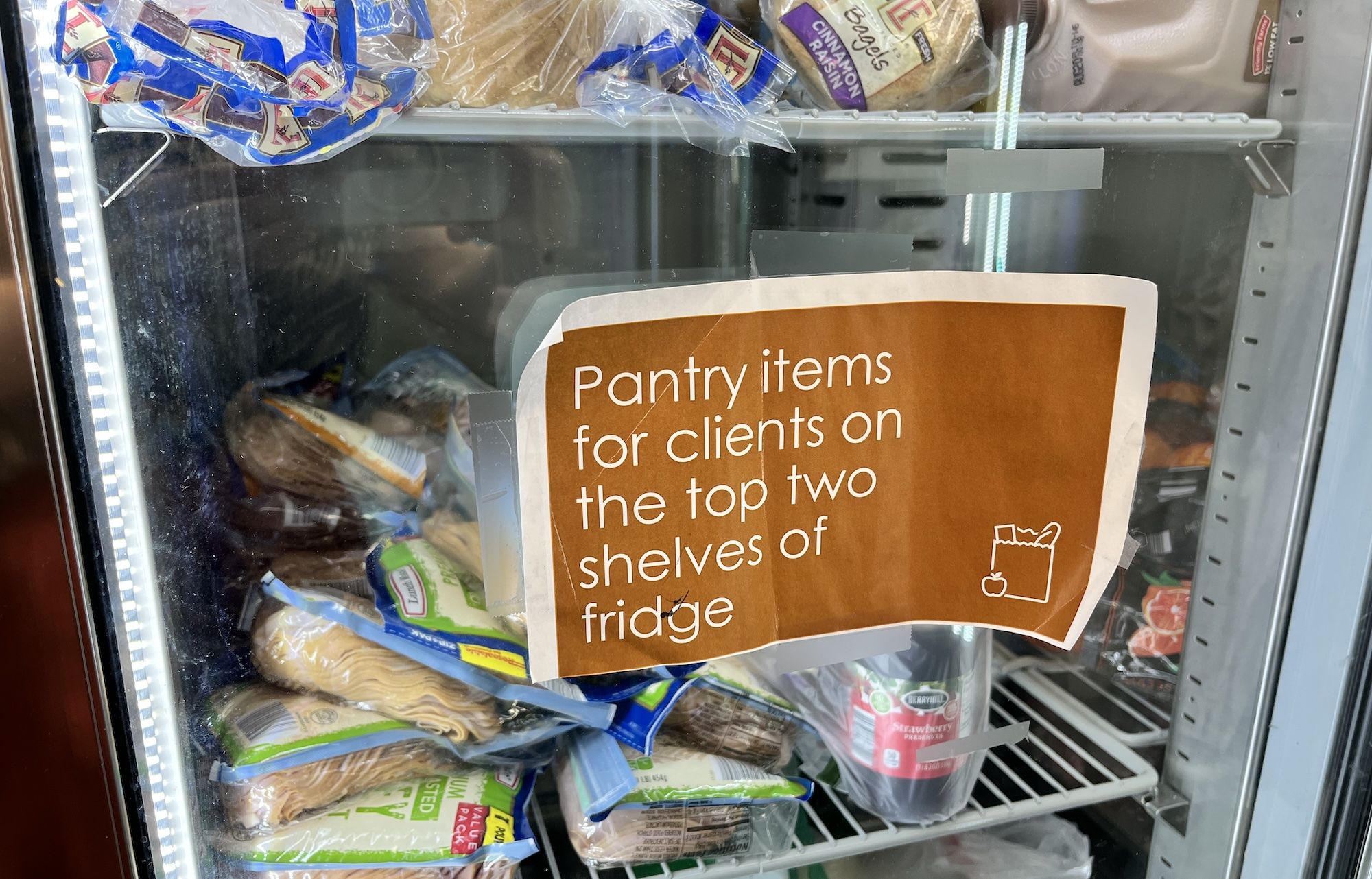 Glass-front fridge with a brown sign that reads "Pantry items for clients on the top two shelves of the fridge", and shelves filled with bread, pastries, and assorted packaged foods.