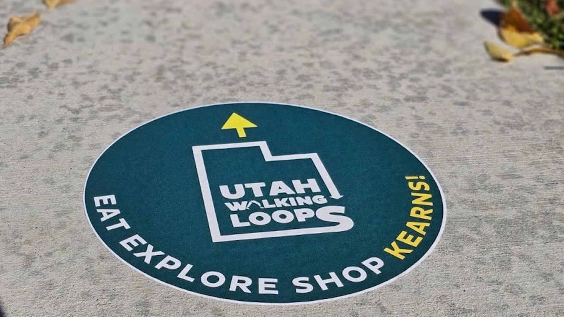 Teal circular sticker on a concrete sidewalk promoting walking loops: "Utah Walking Loops" with a yellow upward arrow, white edge text reading "EAT EXPLORE SHOP LEARN".