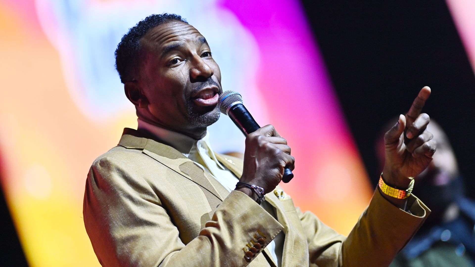 A Black man in a light-tan suit and gold watch points toward the audience while speaking into a microphone in front of a blurry and colorful LED screen.