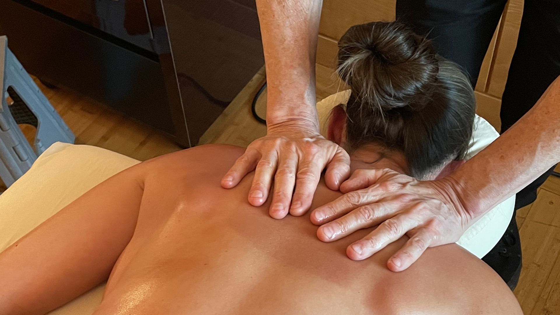 A massage therapist's hands rest on his client's bare shoulders as he prepares to rub CBD massage oil over her back. 