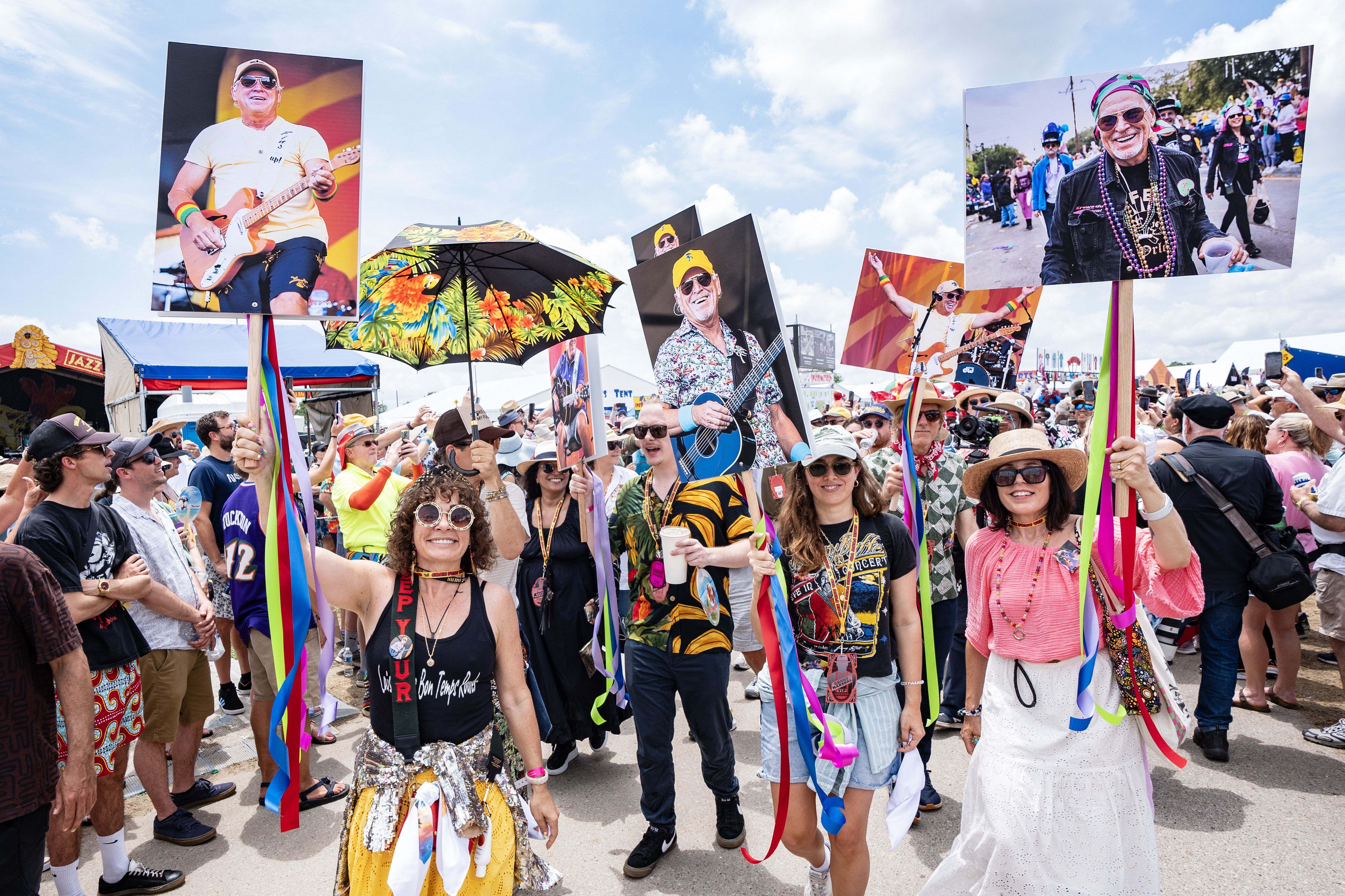 A second-line ambles through a Jazz Fest crowd. Women lead the parade, holding up large photos of Jimmy Buffett while at Jazz Fest in various years.