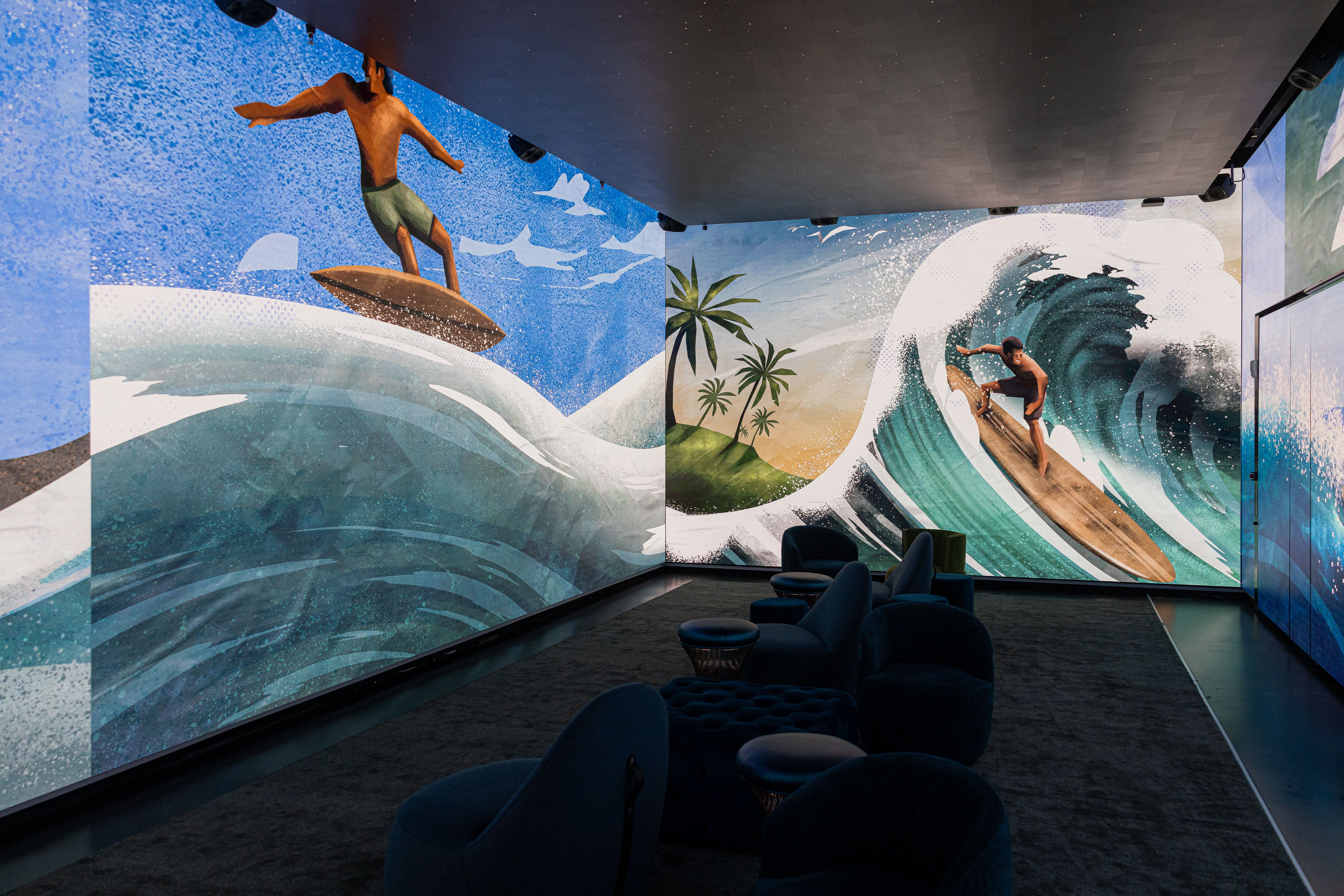Indoor room with large wall murals of surfers riding teal waves under blue sky with white clouds and palm trees, with dark blue chairs and ottomans in foreground.