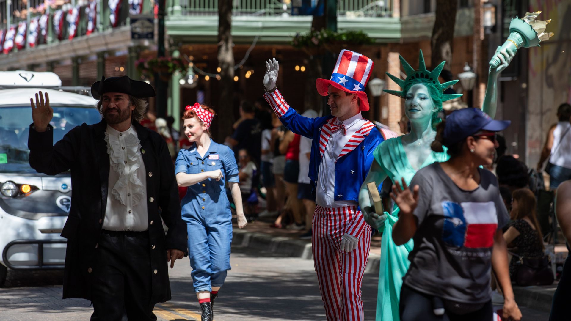 A July Fourth parade on a sunny Houston Street with costumed performers: Uncle Sam in red-and-white striped suit waves; a green Statue of Liberty with a torch; a pirate in black; a woman in denim; patriotic bunting above.