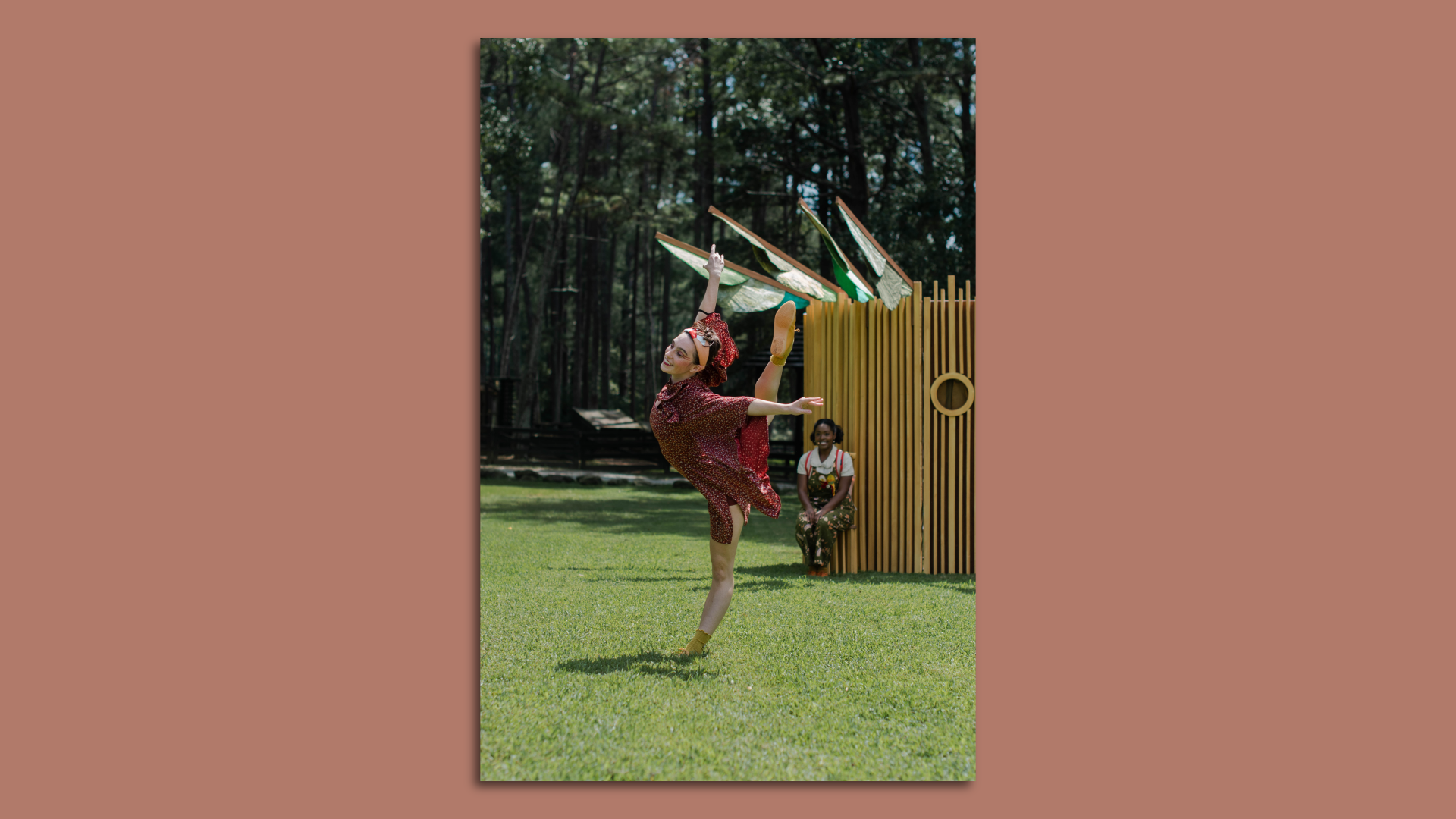 A ballet dancer in red performs outside on a green lawn while a person watches