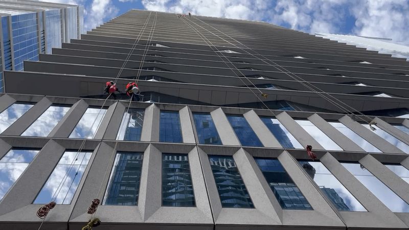 Window washers dangling from a tall building with a blue, cloudy sky above.